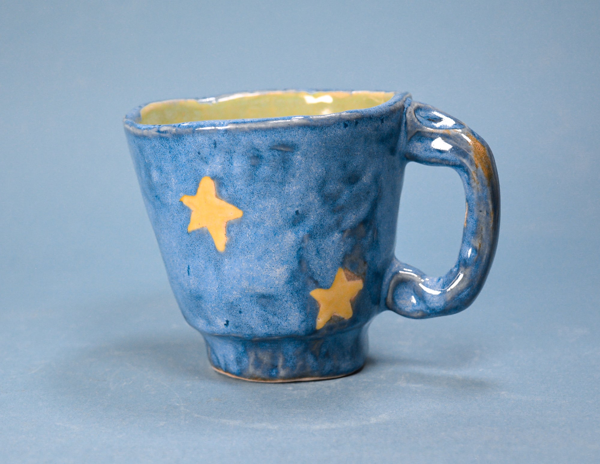 A handmade ceramic mug with a textured blue glaze, decorated with two yellow stars, and a yellow interior, set against a solid blue background.