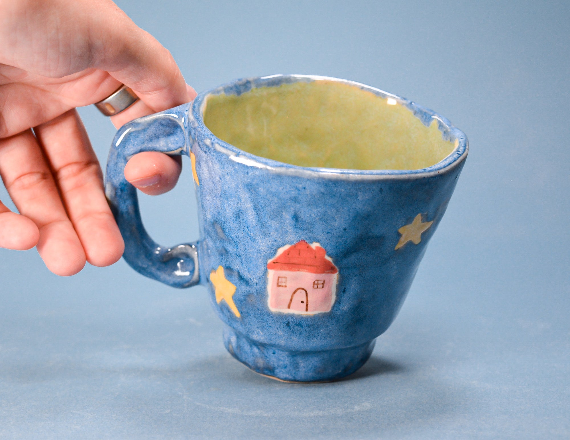 A hand holding a blue ceramic mug with a yellow-green glazed interior, decorated with yellow stars and a small pink house with a red roof.