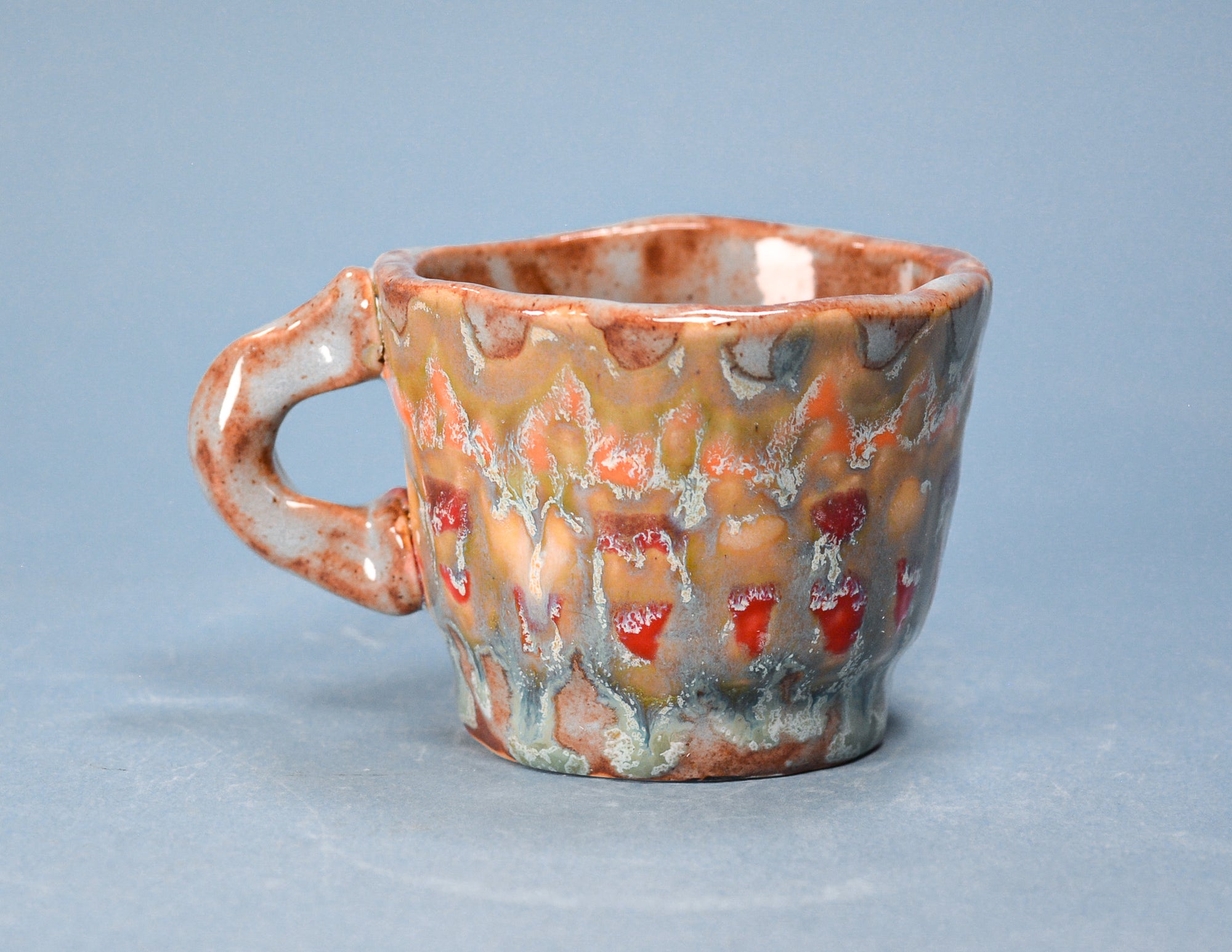 A handmade ceramic coffee cup with a textured, organic pattern in earthy brown, beige, and red glazes, set against a solid blue-gray background.