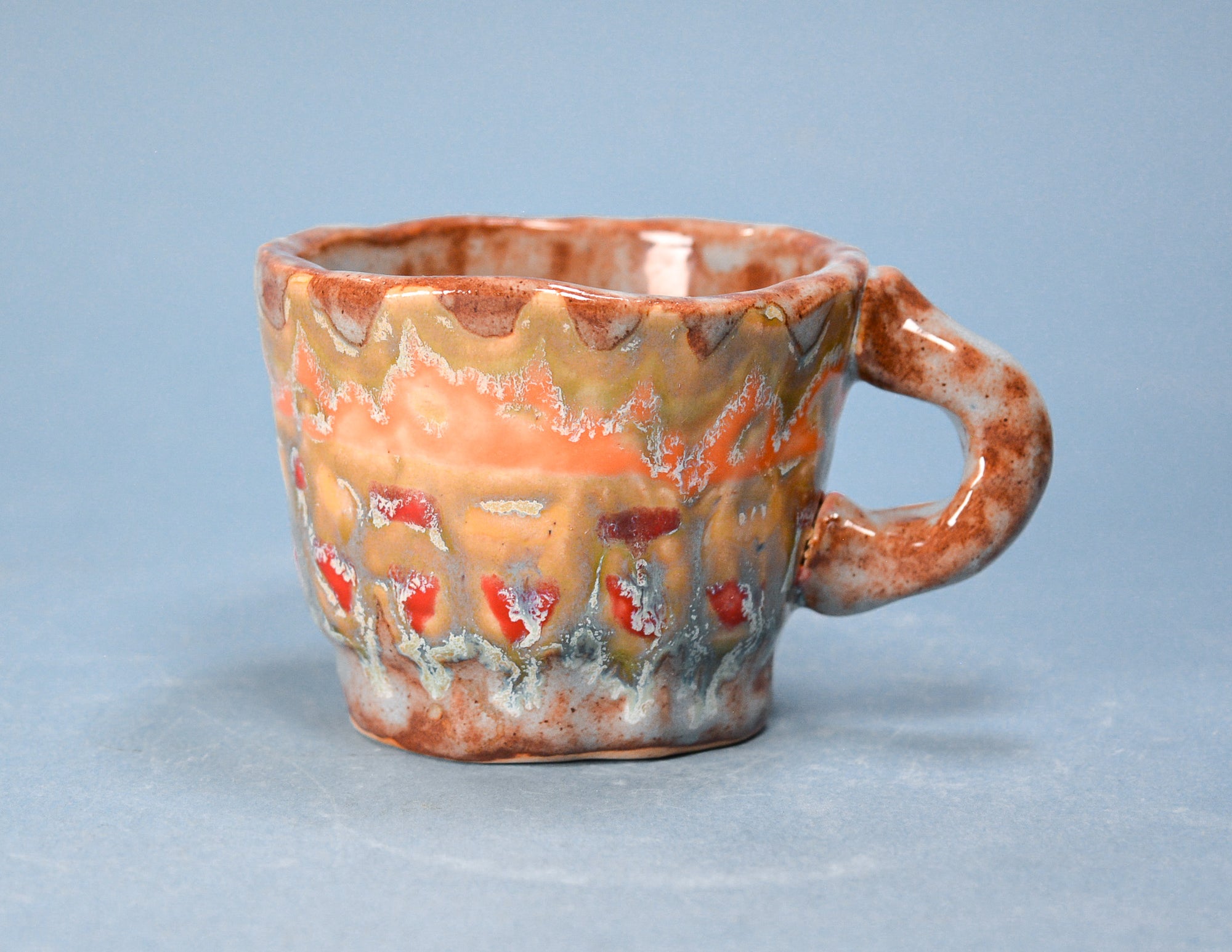 A handmade ceramic cup with a wavy, scalloped rim and a curved handle, decorated with a marbled glaze of orange, brown, beige, and red on a light blue-gray background.