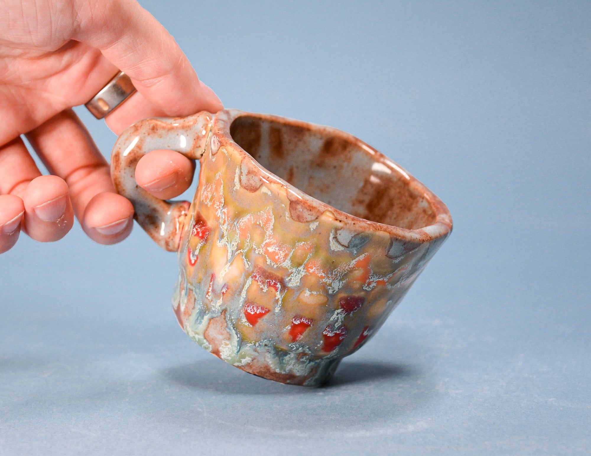 A handmade ceramic mug with a textured, organic glaze pattern in earthy brown, beige, and red, held by a hand against a solid blue-gray background.