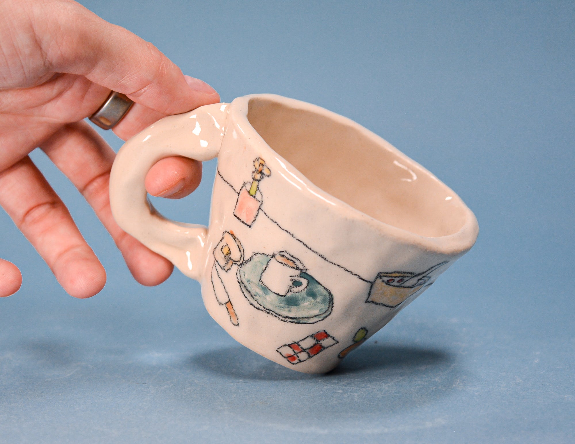 A hand holds a small handmade ceramic mug with a handle, decorated with whimsical illustrations of laundry items on a blue background.