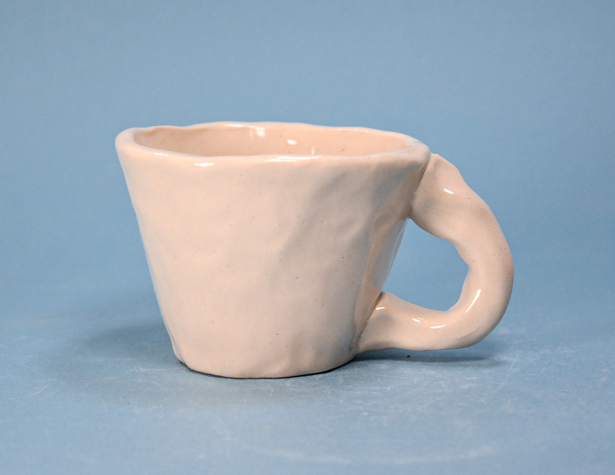 A small handmade ceramic cup with a unique loop handle, finished in a matte pale pink glaze, set against a solid blue background.