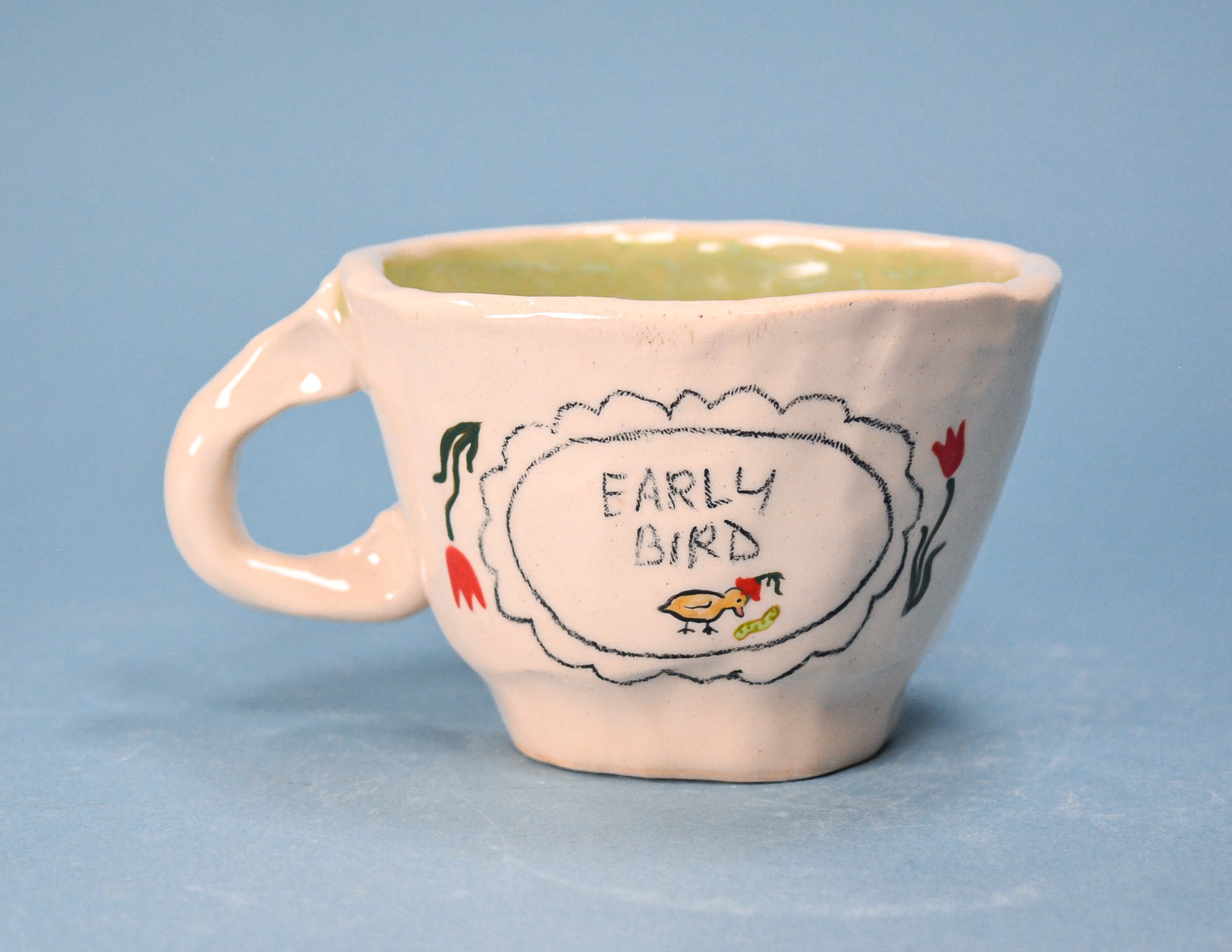A small ceramic coffee cup with a pale pink exterior, a light green interior, and a hand-drawn style label that reads 'EARLY BIRD' with a bird illustration. The cup is photographed against a solid light blue background.