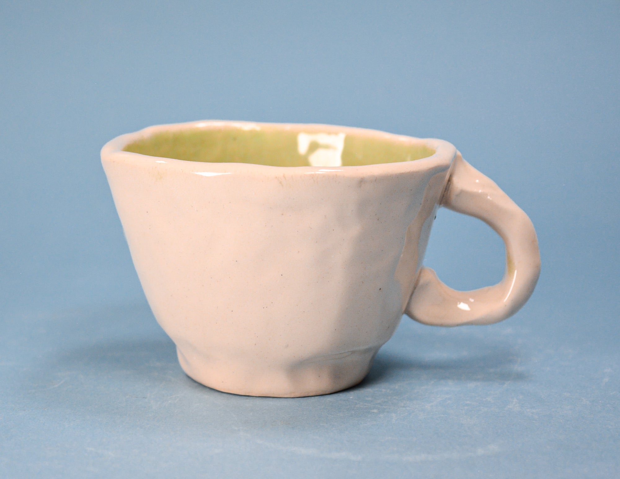 A handmade ceramic cup with a textured, matte off-white exterior and a glossy light green interior, set against a solid blue background.