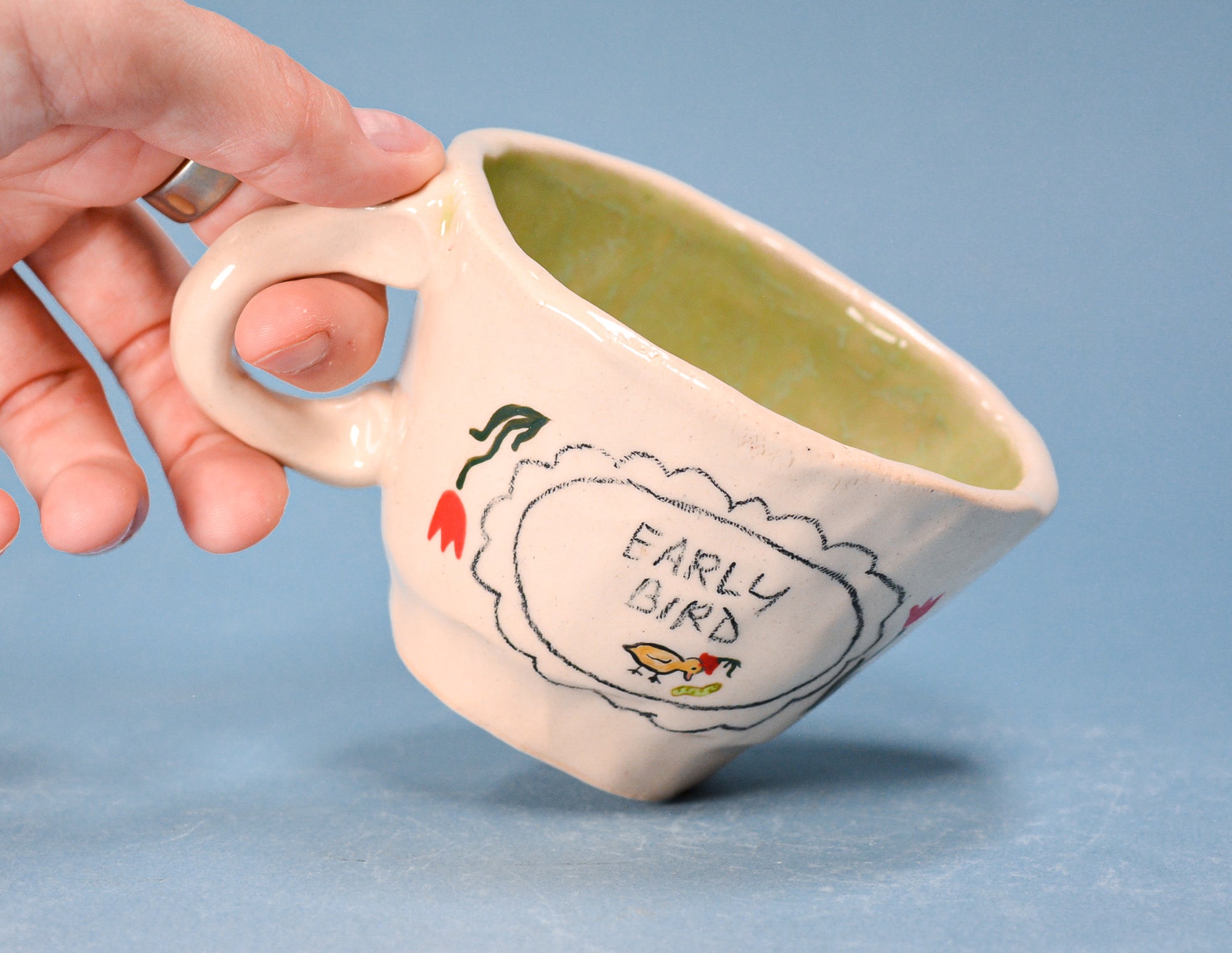 A hand holding a handmade ceramic coffee mug with a light pink exterior, green glazed interior, and a hand-painted design that reads 'EARLY BIRD' with a bird and tulip on a blue background.