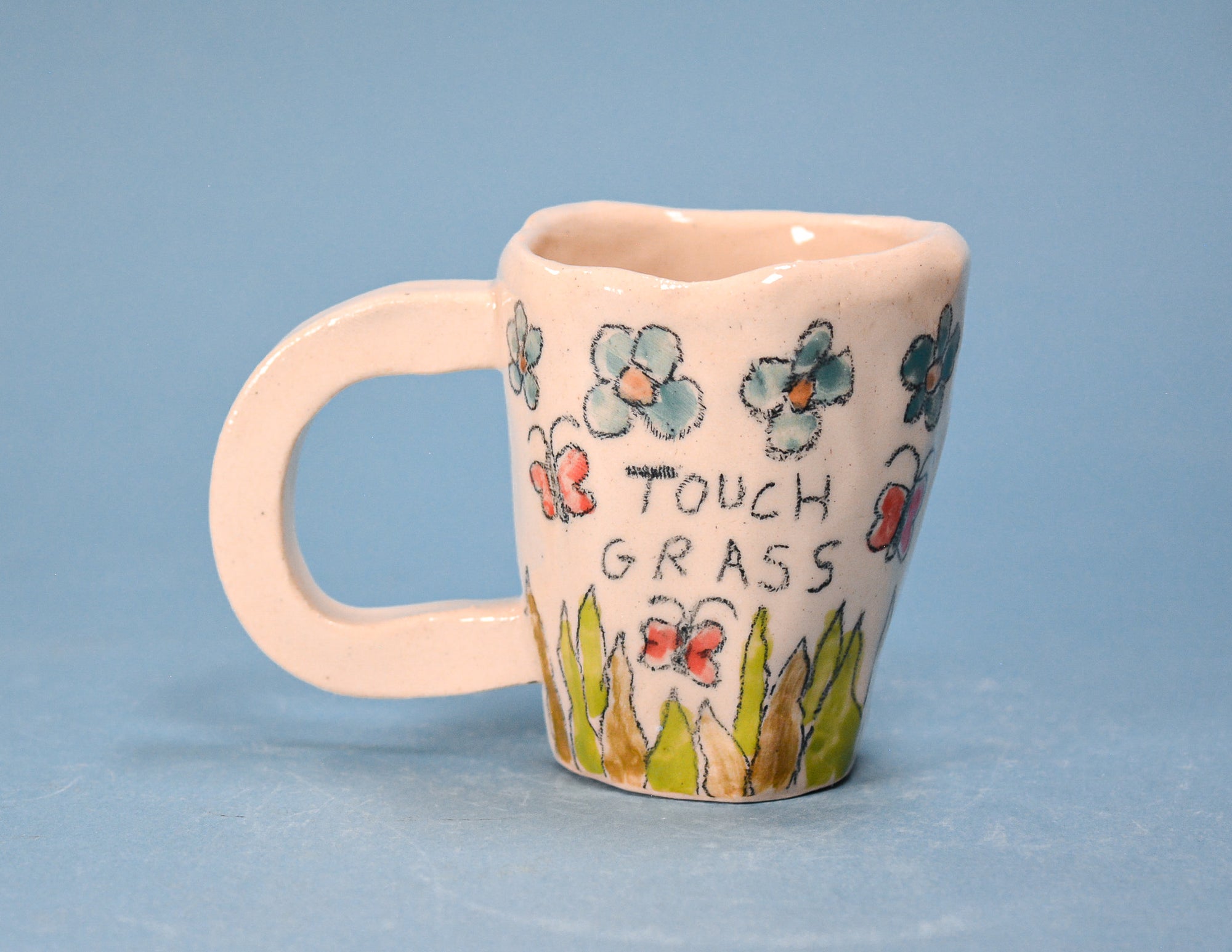 A small ceramic mug with a pale pink handle and body, decorated with blue flowers, red ladybugs, green grass, and the words 'TOUCH GRASS' in black lettering, set against a solid blue background.