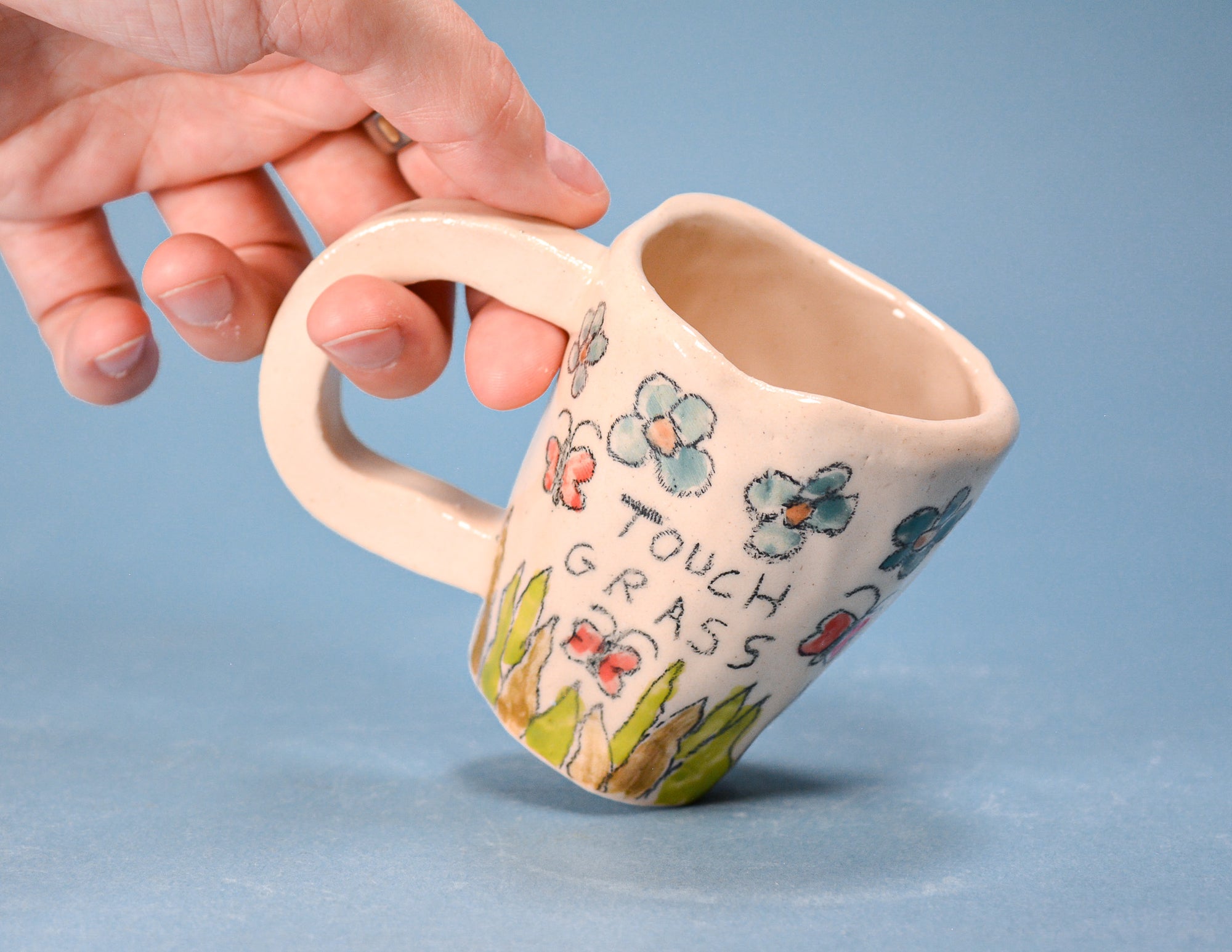 A hand holds a small ceramic mug against a solid blue background. The mug is cream-colored with hand-painted flowers, butterflies, and the words 'TOUCH GRASS' in black.