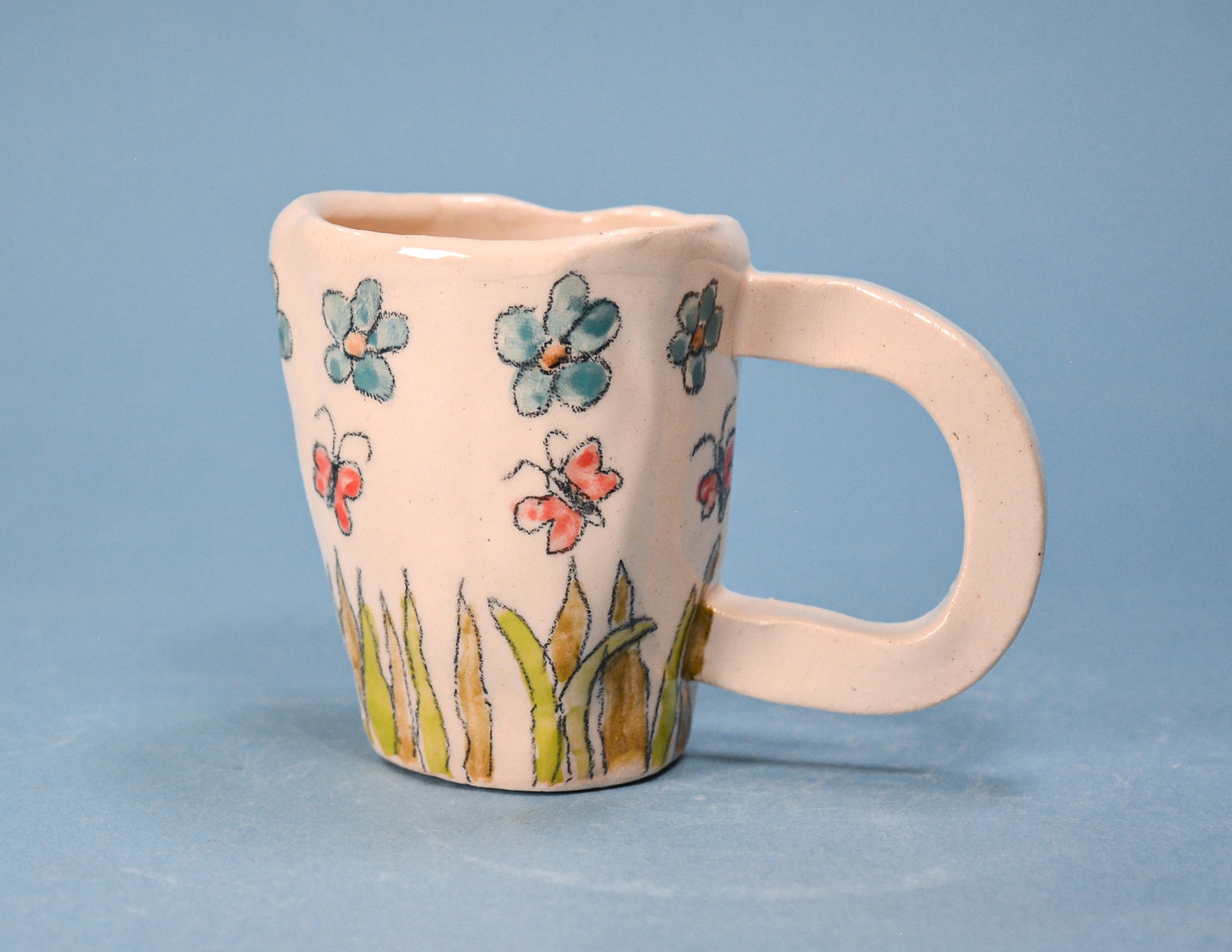 A small ceramic mug with a rounded handle, featuring a light pinkish-beige glaze decorated with hand-painted blue, red, and green flowers and grass, set against a solid blue background.