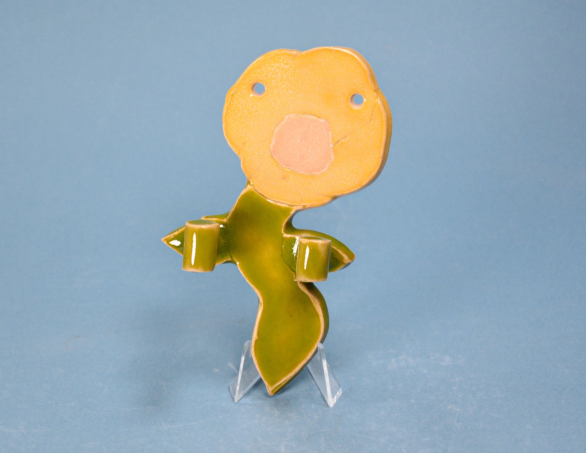 A small decorative figurine or sculpture shaped like a stylized plant or character, with a round yellow-orange head, green body, and clear plastic display stand, photographed against a plain blue background.