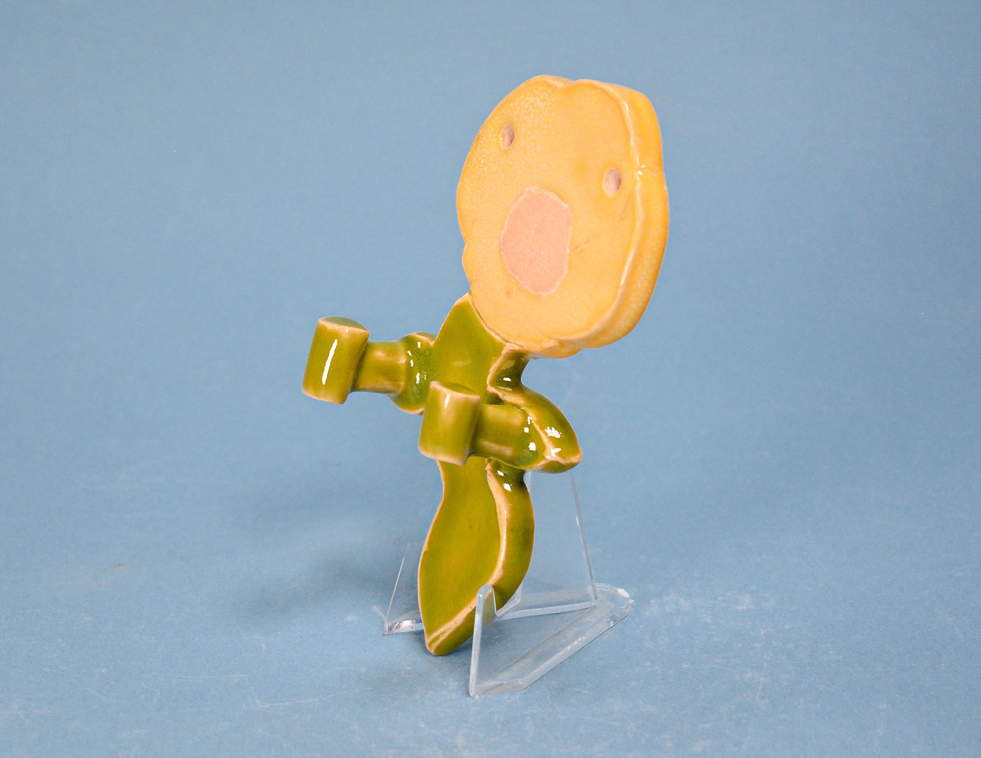 A small glossy figurine of a stylized flower or plant with a round yellow-orange bloom, olive-green stem and leaves, standing on a clear plastic display stand against a light blue background.
