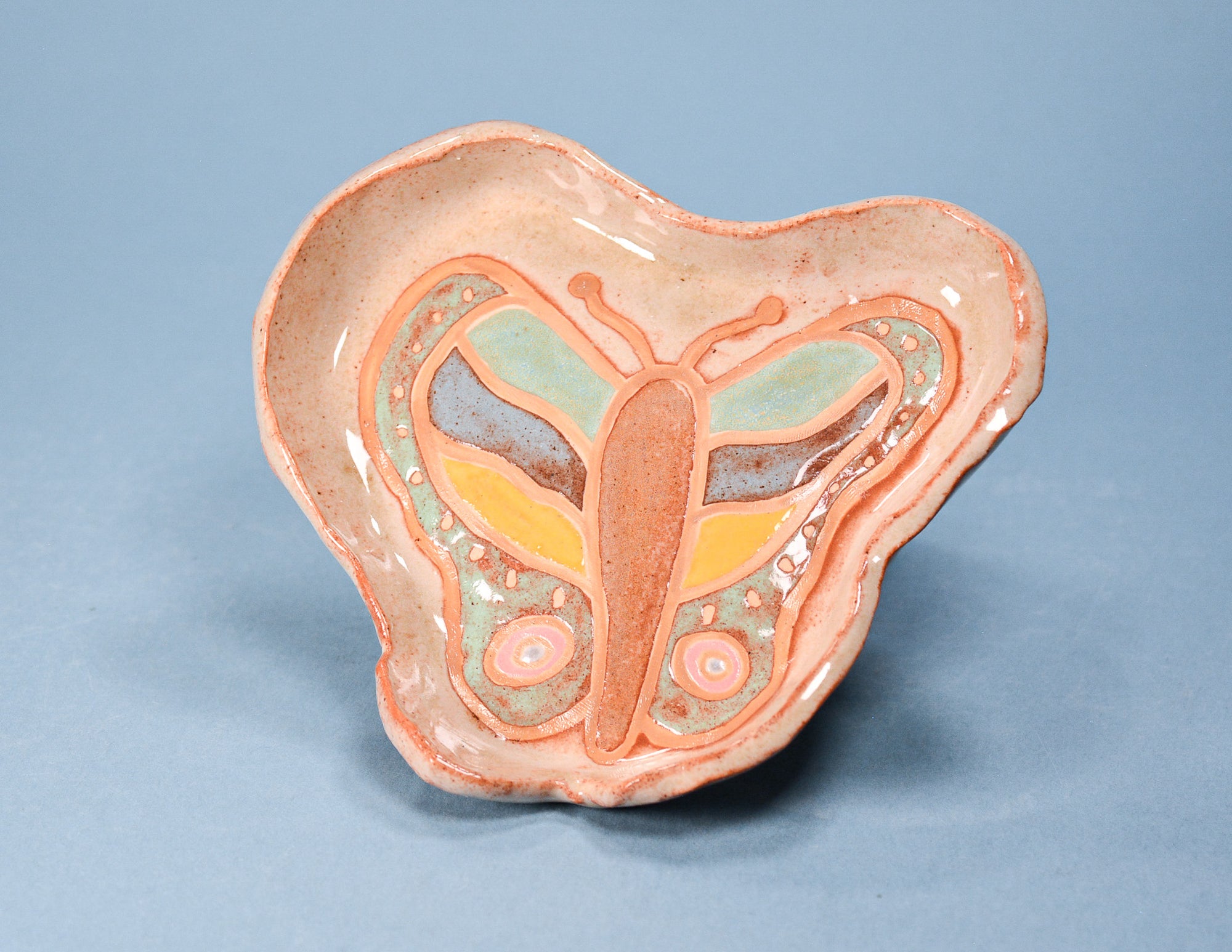 A handmade ceramic dish shaped like a butterfly, featuring a peach-colored glaze with a painted butterfly design in yellow, light blue, and gray on a solid blue background.