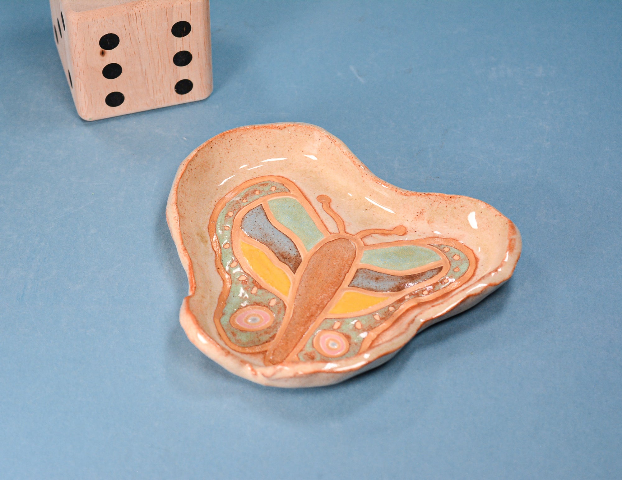 A handmade ceramic butterfly dish with a glossy peach and orange glaze, featuring a painted butterfly design in yellow, green, and blue. A wooden die is visible in the top left corner for scale, set against a solid blue background.