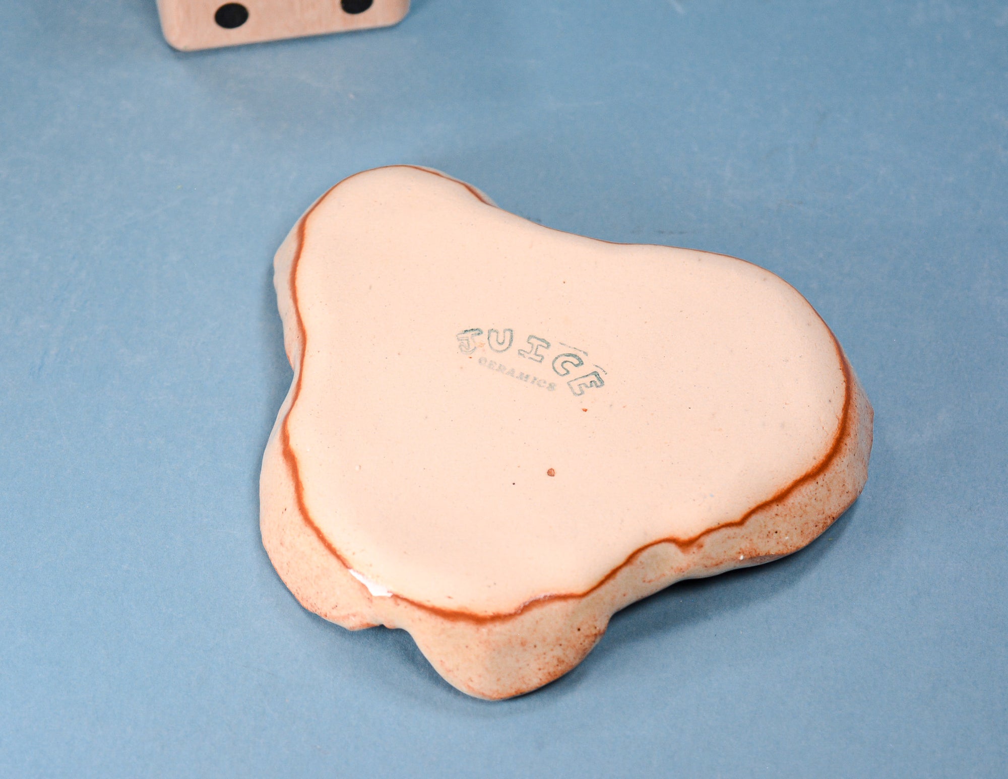 A bone-shaped ceramic coaster or tile with a glossy pale pink glaze and a thin terracotta rim, featuring the debossed brand name 'FUSE' in the center, set against a textured light blue background.