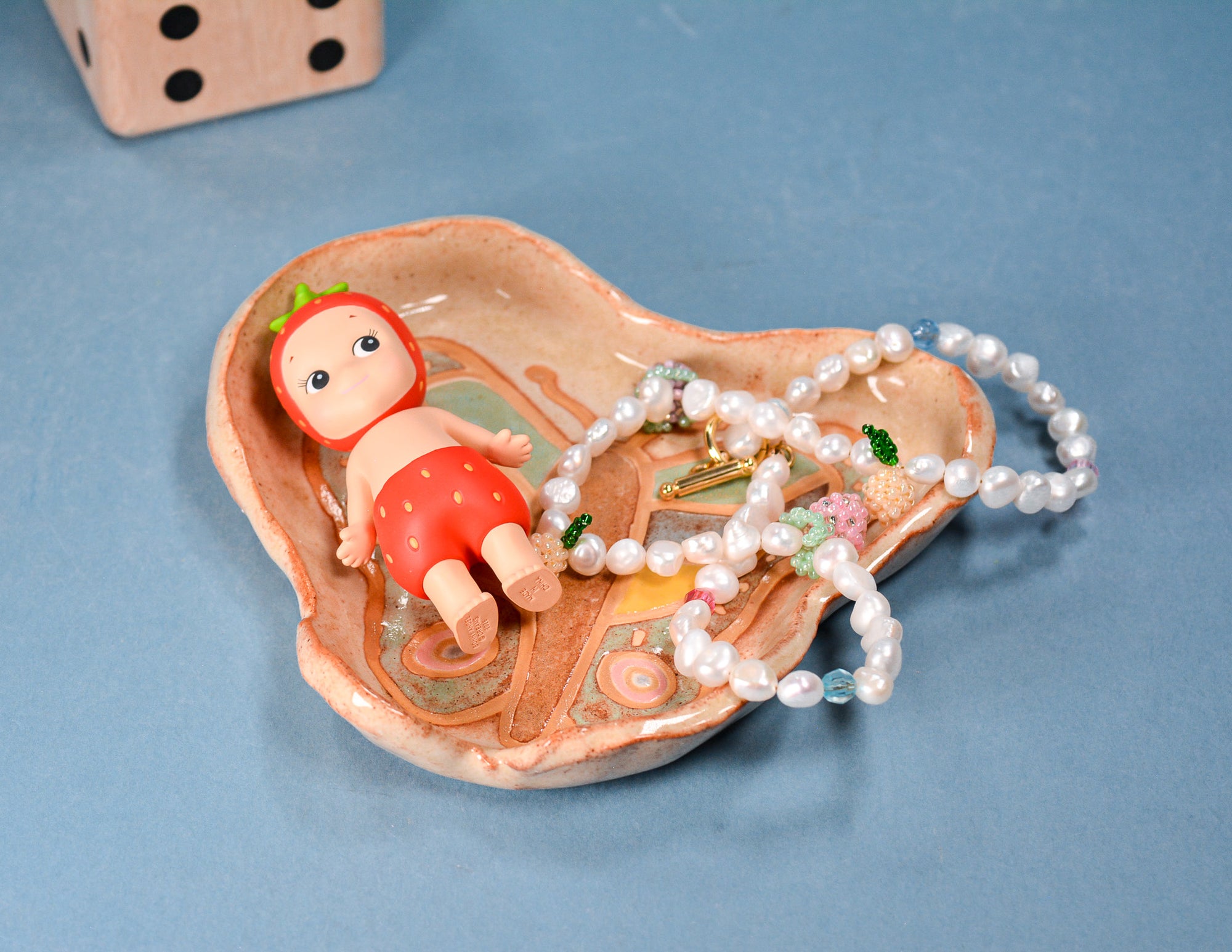 A heart-shaped ceramic jewelry tray with a painted floral design, holding a strand of white pearl beads and a small doll accessory, set against a blue background.
