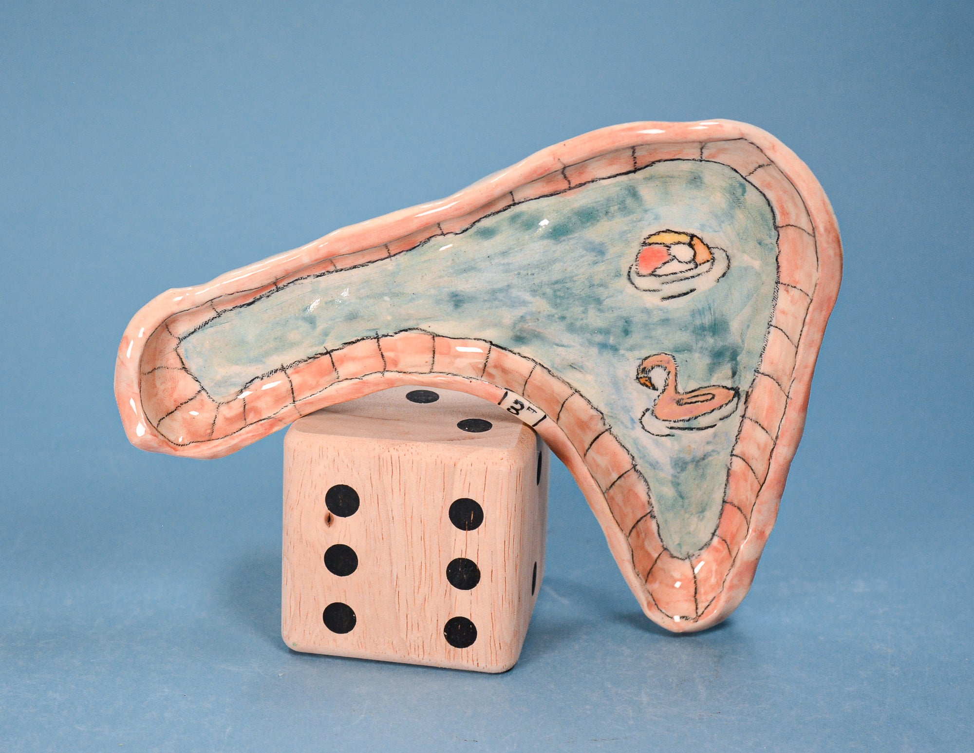 A ceramic novelty pool shaped like a curved swimming pool with a peach-colored rim and light blue water surface, featuring painted swan illustrations. The table is shown balancing on a large wooden die against a solid blue background.