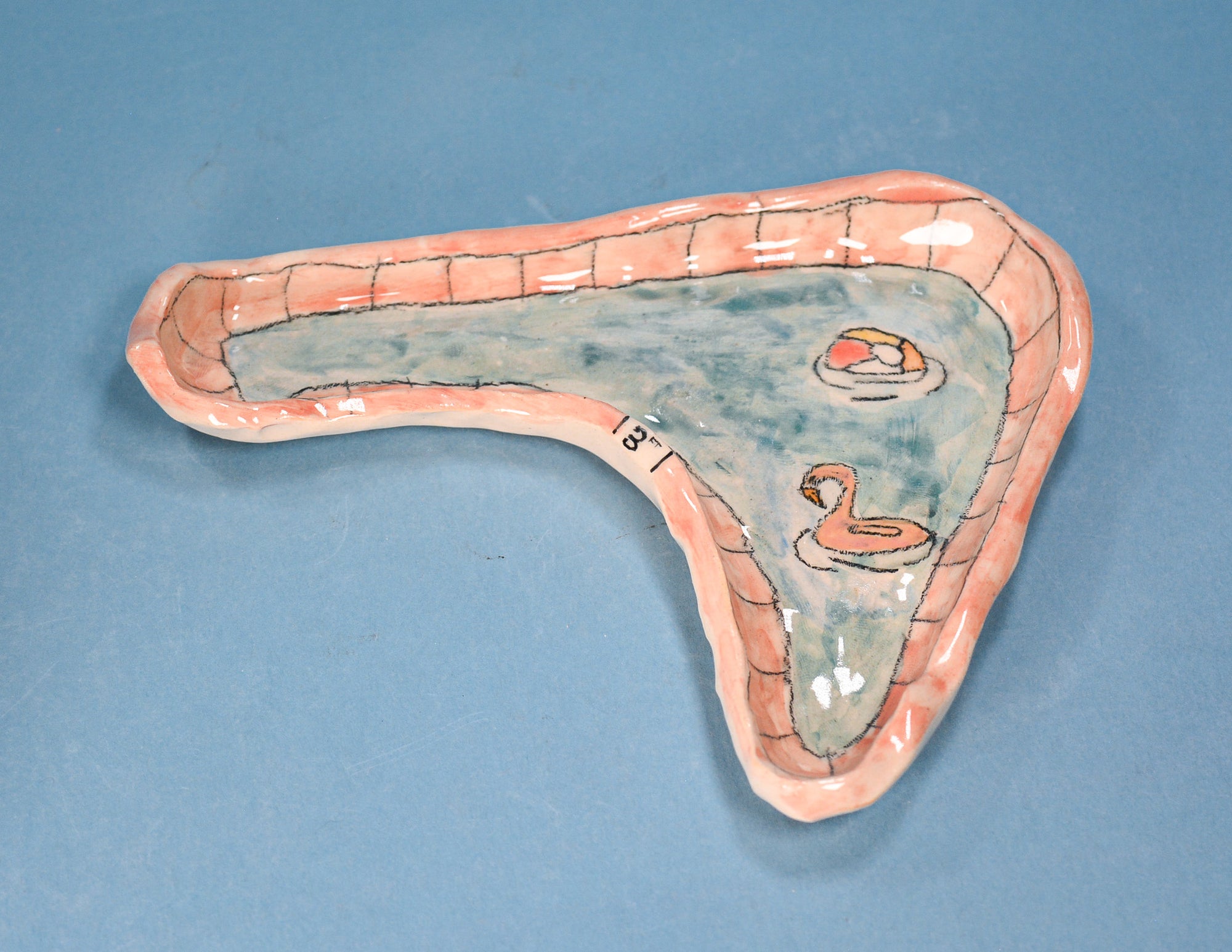 A whimsical, hand-painted ceramic pool tray shaped like a swimming pool with a peach-pink border, blue-green water, and illustrated swans on a blue background.