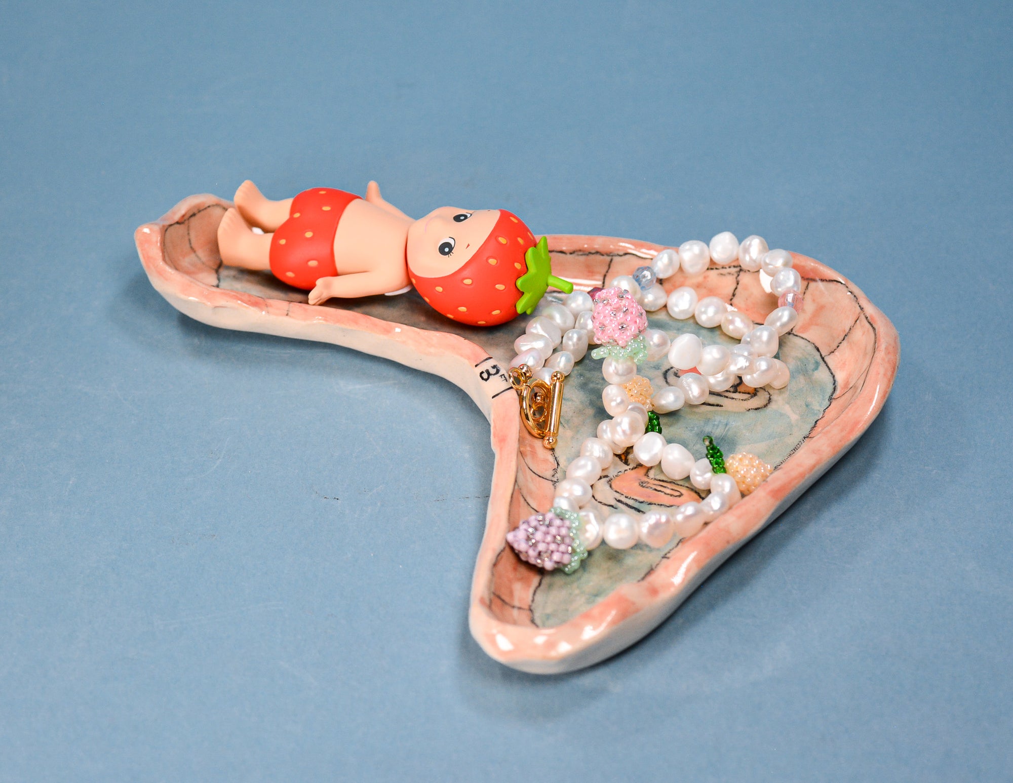 A ceramic heart-shaped jewelry organizer tray with a strawberry-themed design, featuring a small figurine of a baby in red strawberry shorts and a strawberry hat, holding pearl necklaces and bracelets on a blue background.