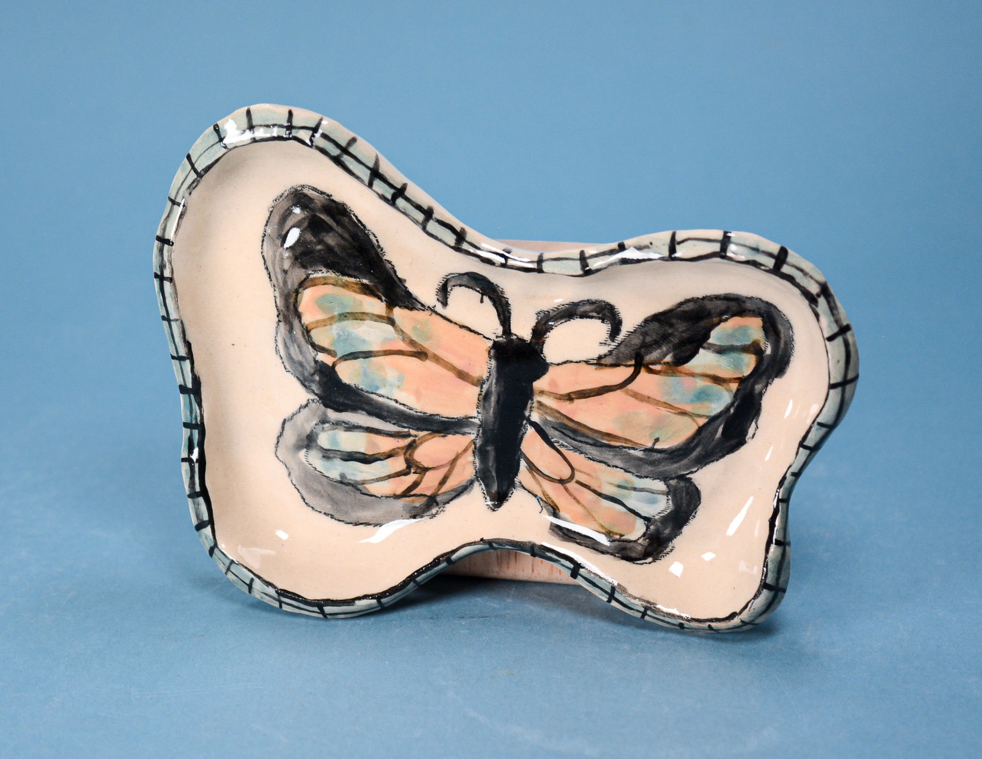 A butterfly-shaped ceramic dish with a hand-painted monarch butterfly design on a light pink background, set against a solid blue backdrop.