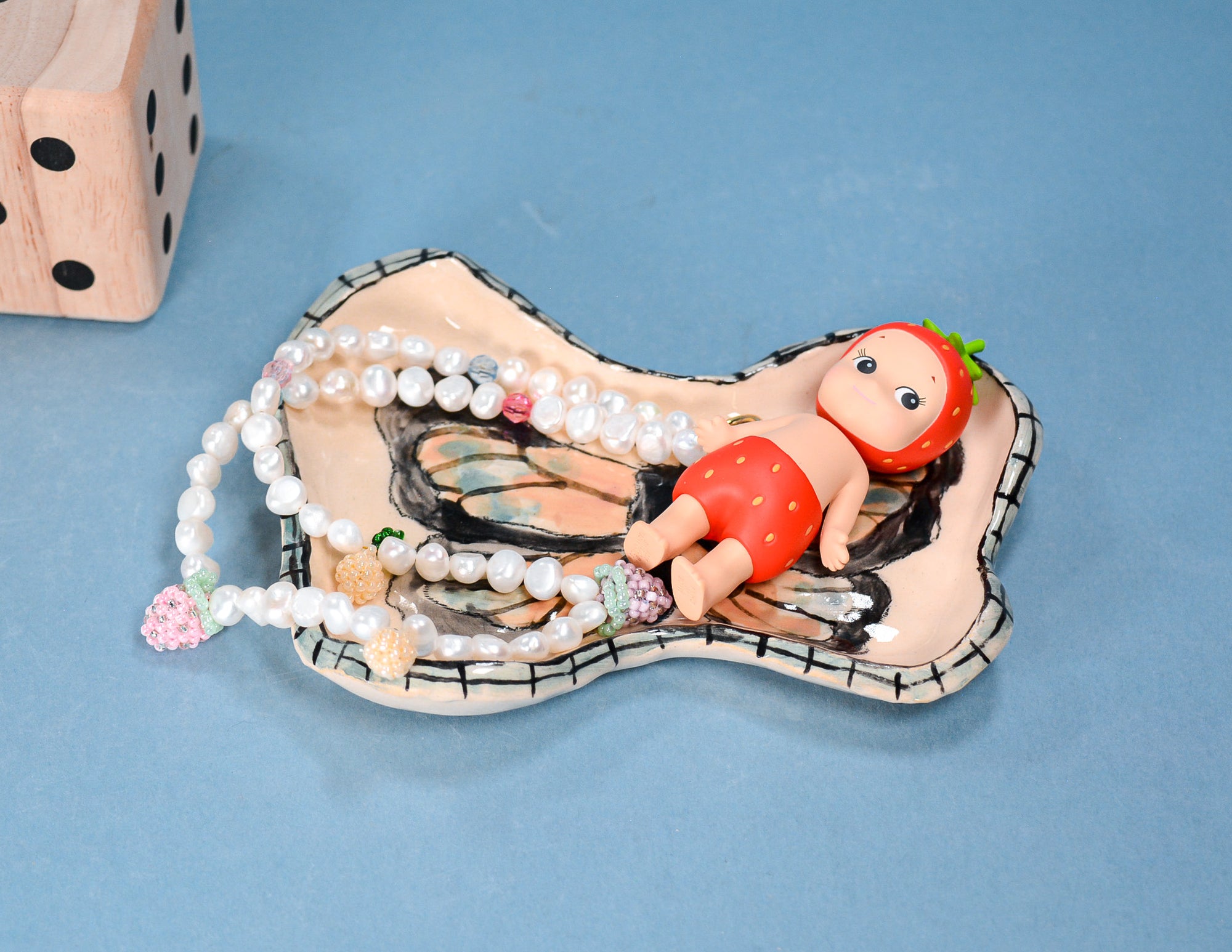 A ceramic butterfly-shaped dish with a painted butterfly wing design, holding a white pearl strand and a small strawberry-themed doll, set against a blue background with a wooden die partially visible in the corner.