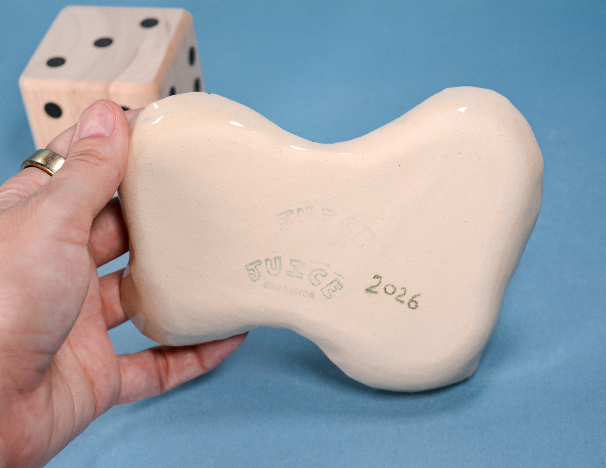 A hand holds a glossy bone-shaped ceramic object against a blue background, with a large wooden die visible in the upper left. The object appears to be the product, with embossed text reading 'FUSGE' and '2026' on the bottom.