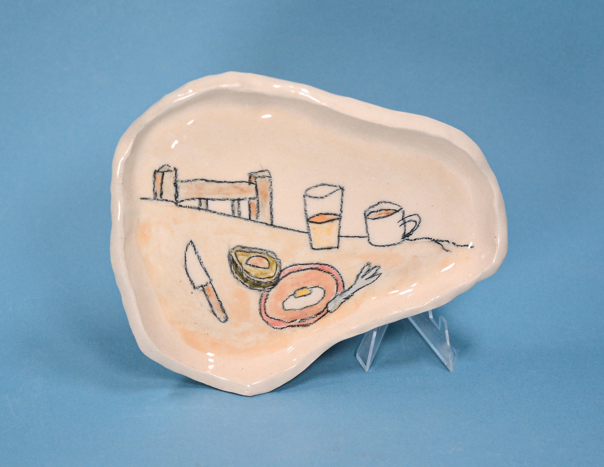 An organic-shaped ceramic serving platter with a hand-painted breakfast scene, displayed on a small clear stand against a solid blue background.