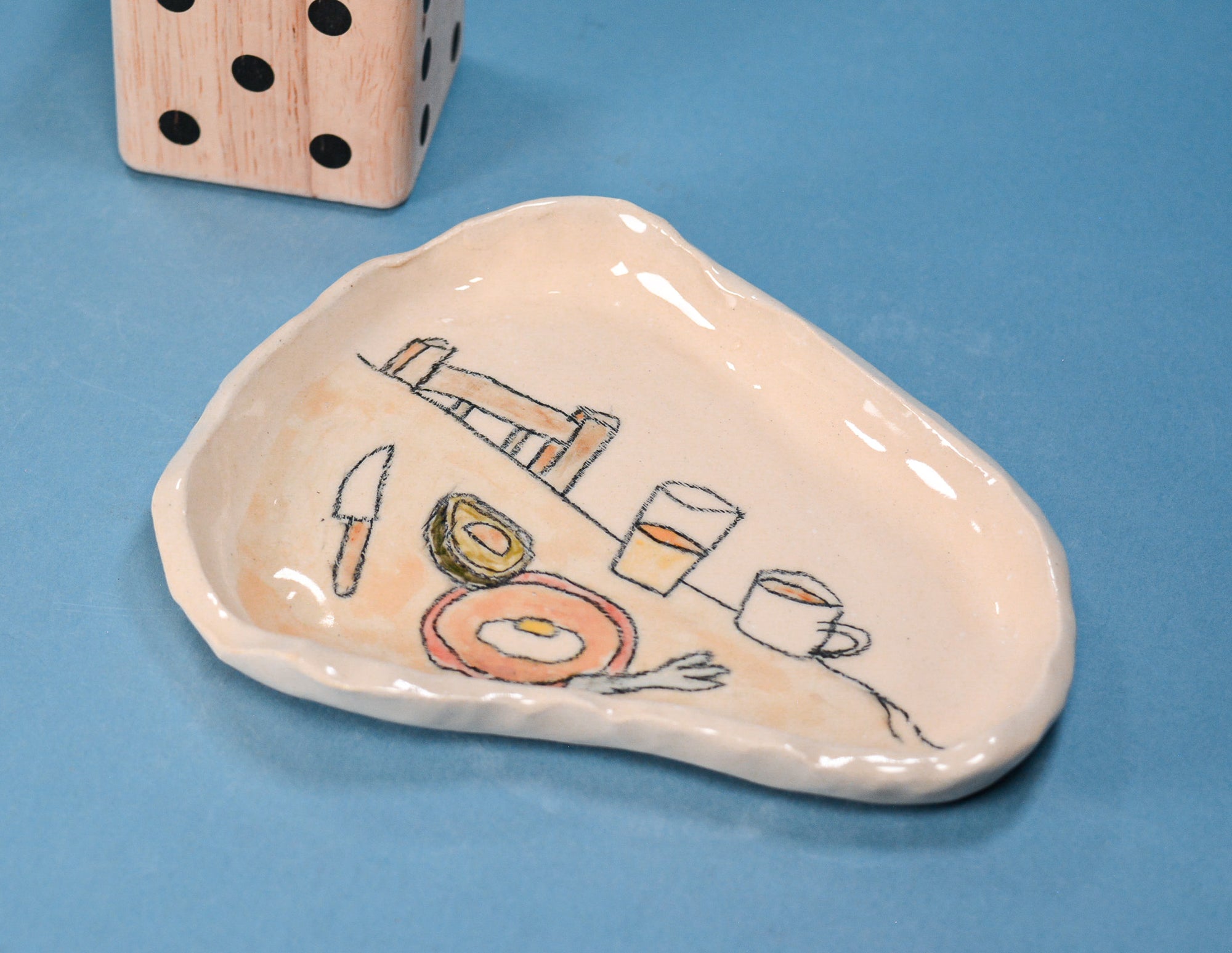 An irregularly shaped cream-colored ceramic dish with a hand-painted breakfast illustration, set against a solid blue background with a wooden die partially visible in the top left corner.