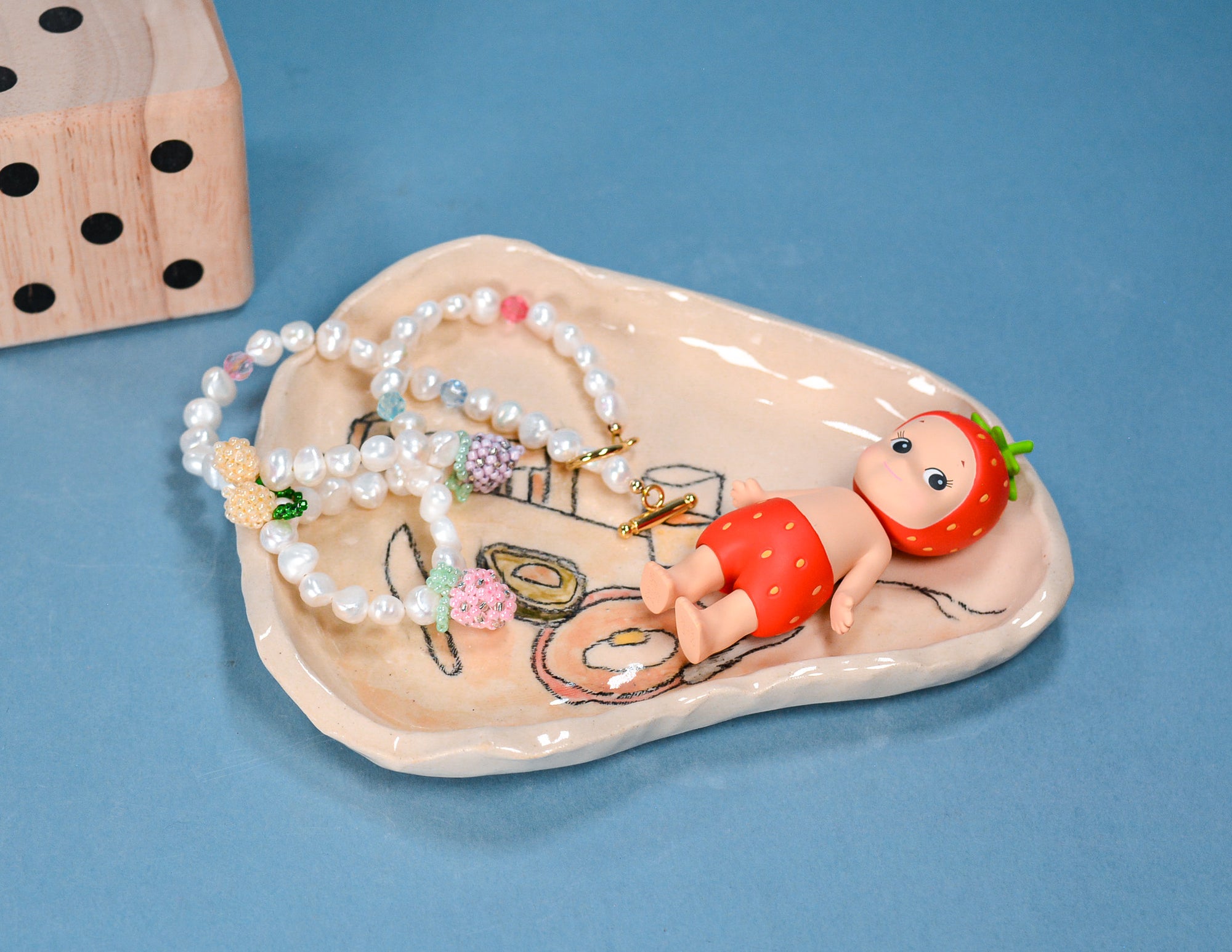 A ceramic jewelry tray with a whimsical strawberry-themed design, holding a beaded necklace, set against a solid blue background with a wooden die prop in the corner.