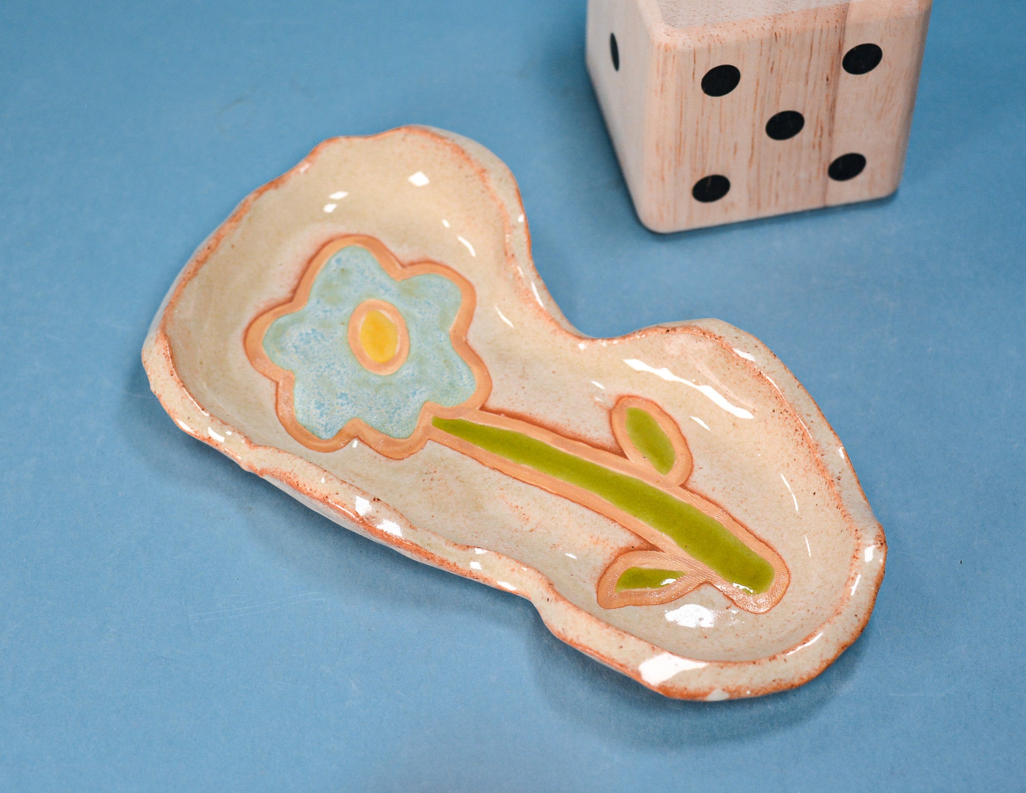 A handmade ceramic dish with a hand-painted flower design, featuring a light blue flower with a yellow center and a green stem on a peach and beige glazed base, shown next to a wooden die on a blue background.