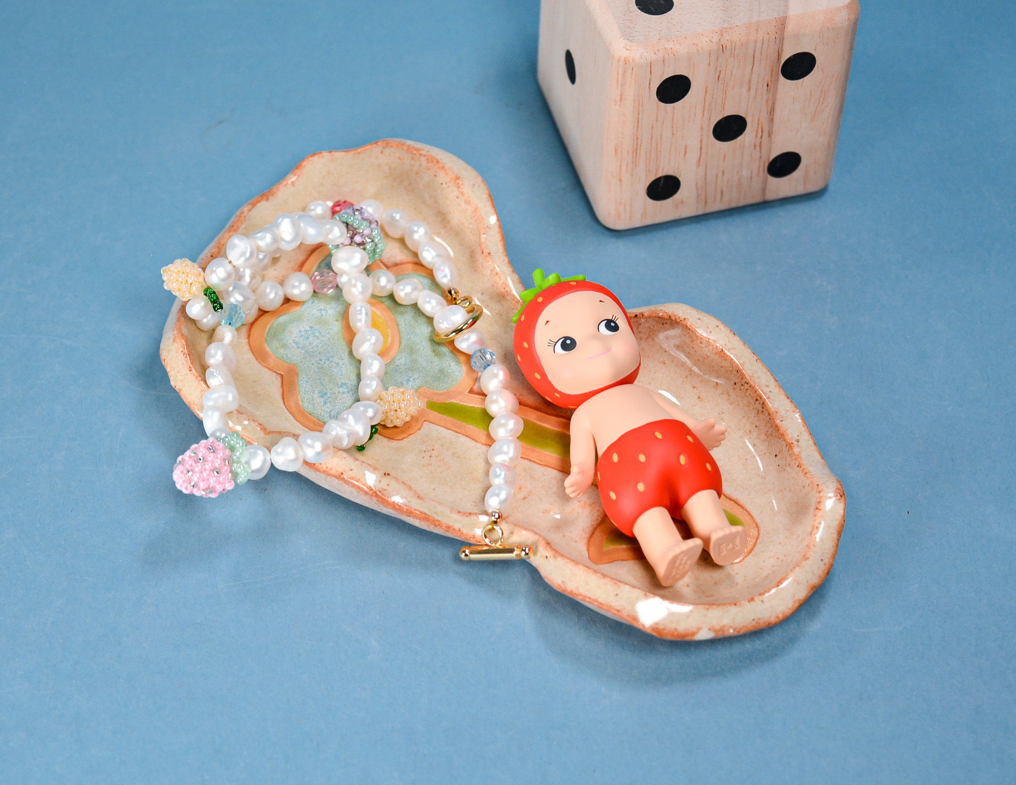 A ceramic strawberry-shaped jewelry holder on a blue background, containing a pearl necklace and a small doll with a strawberry hat and red shorts, with a wooden die in the background.
