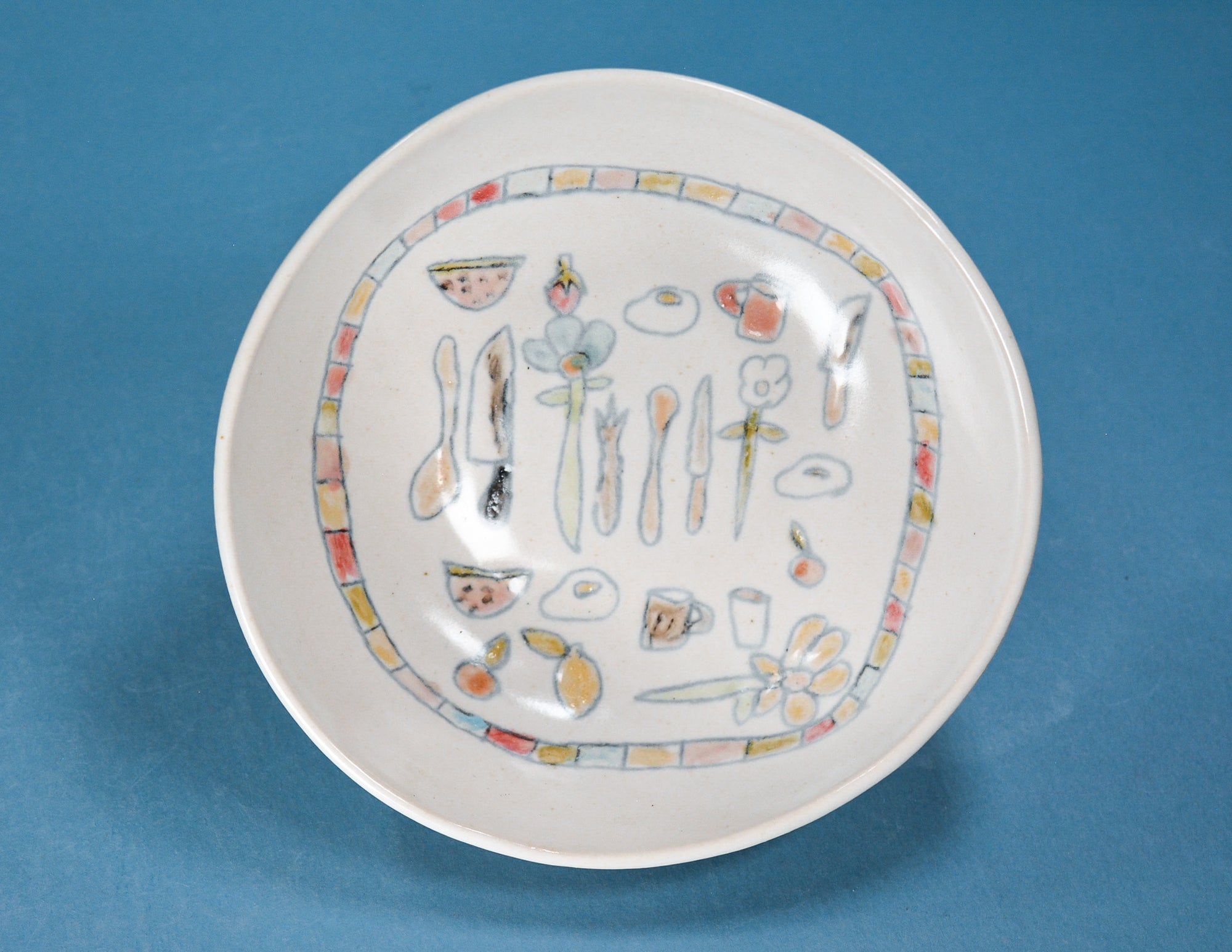A white ceramic plate with a colorful hand-painted illustration of kitchen utensils, flowers, and food items inside a decorative border, set against a solid blue background.