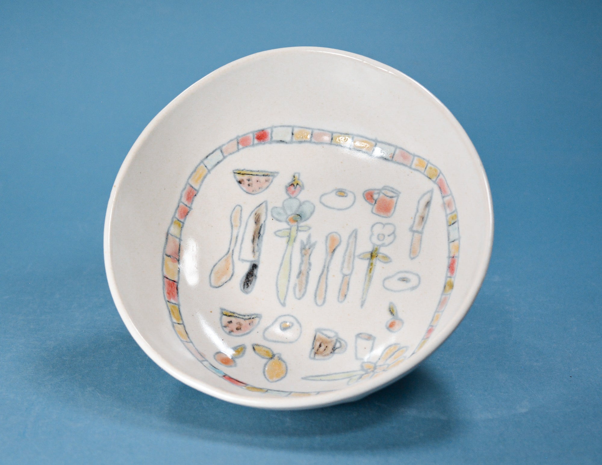 A white ceramic bowl with a colorful hand-painted illustration of kitchen utensils and food items inside, set against a solid blue background.