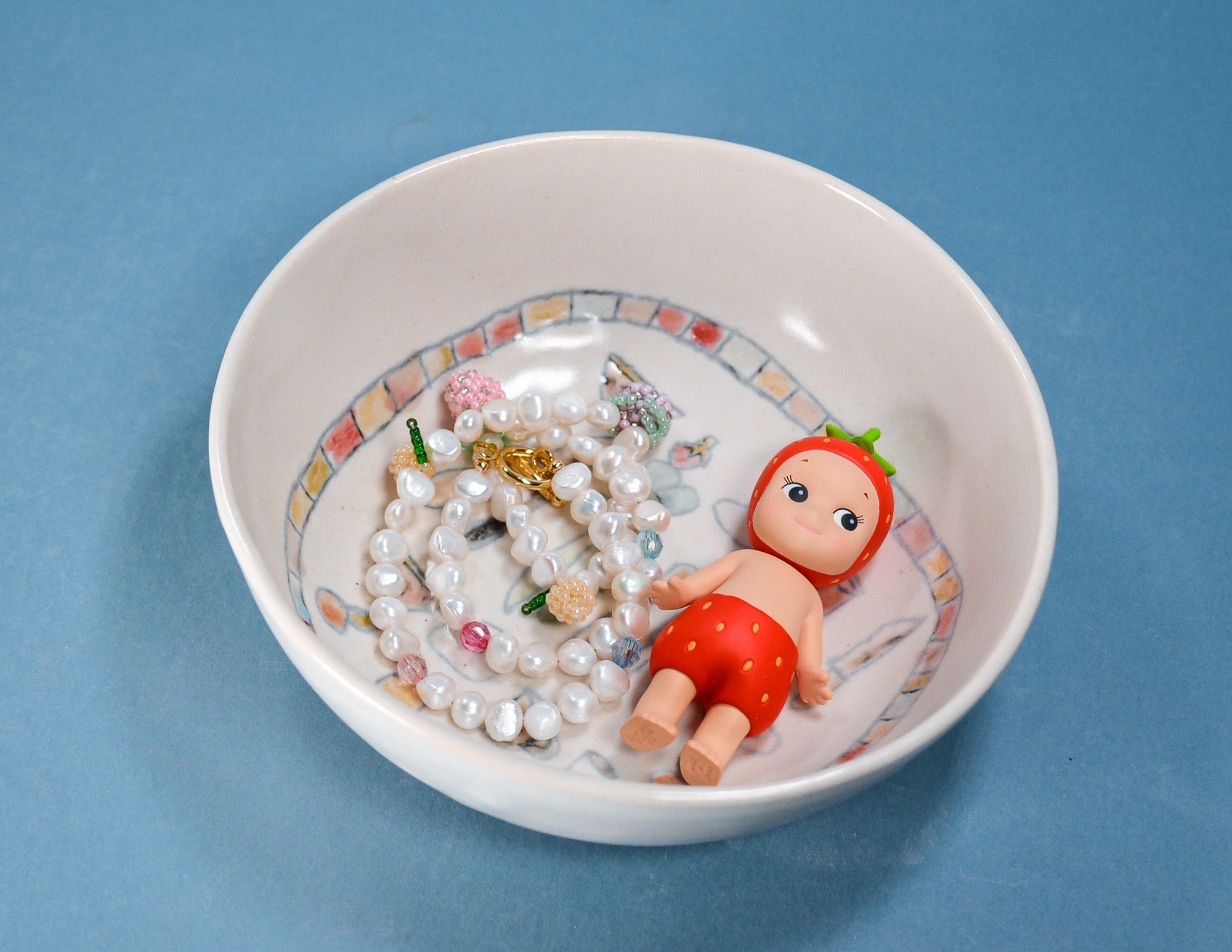 A white ceramic bowl with a colorful patterned rim, containing a pearl necklace and a small doll dressed in red, set against a solid blue background.