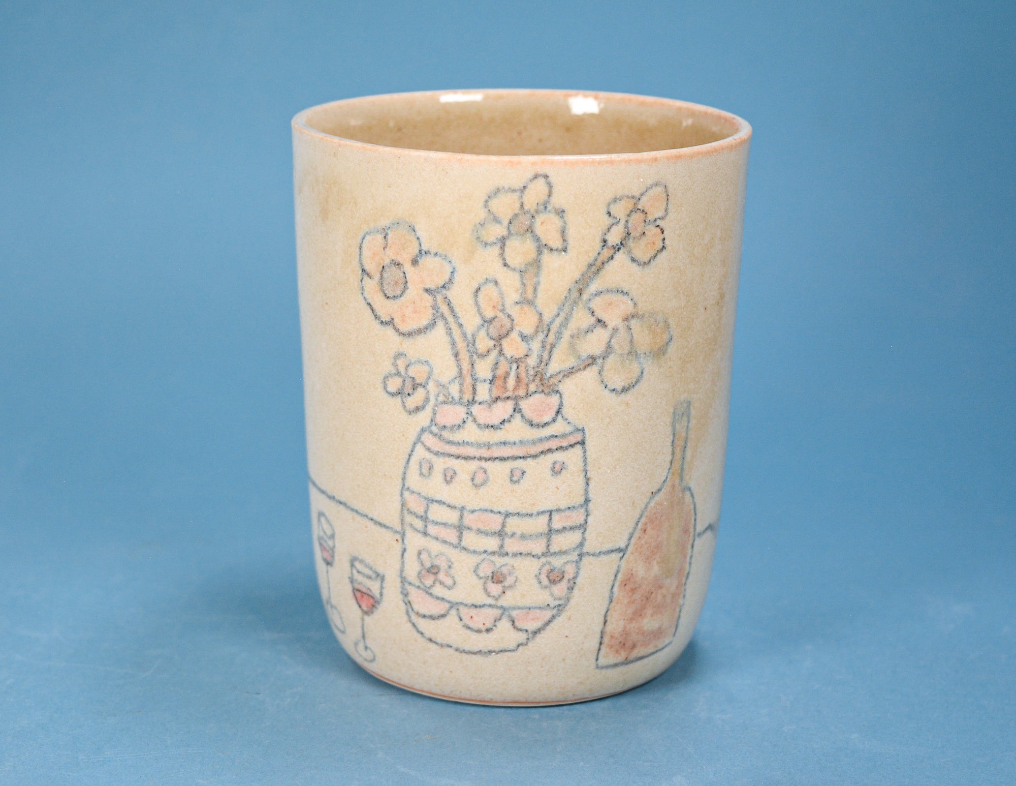 A handmade ceramic cup with a beige base, decorated with a hand-drawn style illustration of a flower vase, flowers, a wine glass, and a bottle, set against a solid blue background.