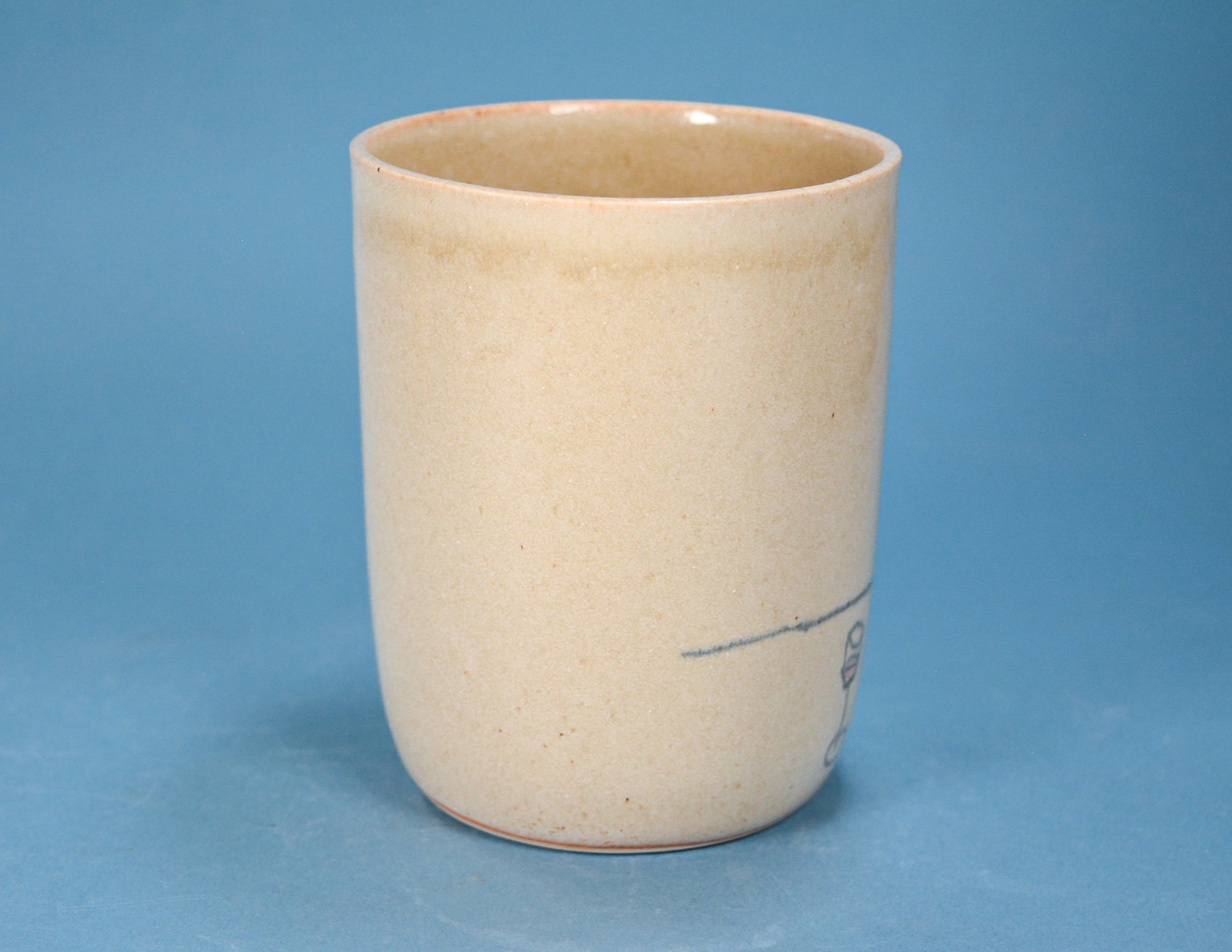 A handmade ceramic cup with a warm beige speckled glaze and a small dark blue line design near the base, set against a solid blue background.