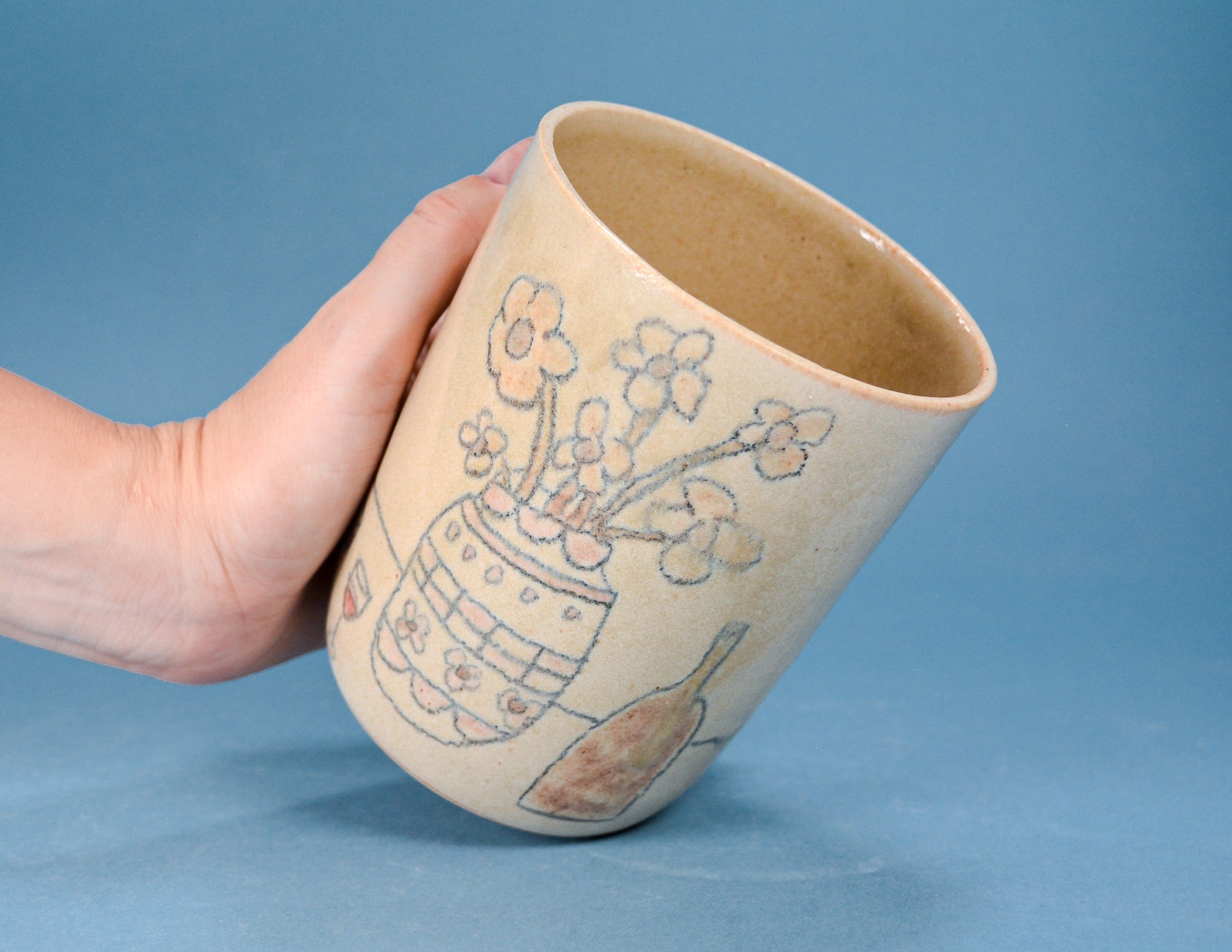 A hand holding a handmade stoneware cup or vase with a floral and botanical illustration, set against a solid blue background.