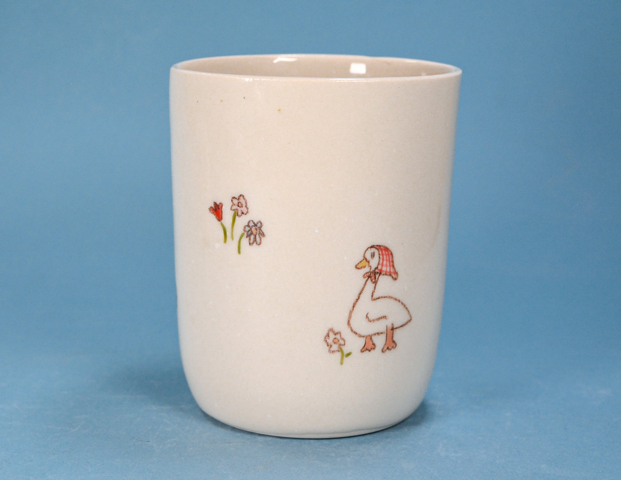 A vintage cream-colored ceramic cup featuring a hand-painted illustration of a duck wearing a red hat and small flowers, set against a solid blue background.