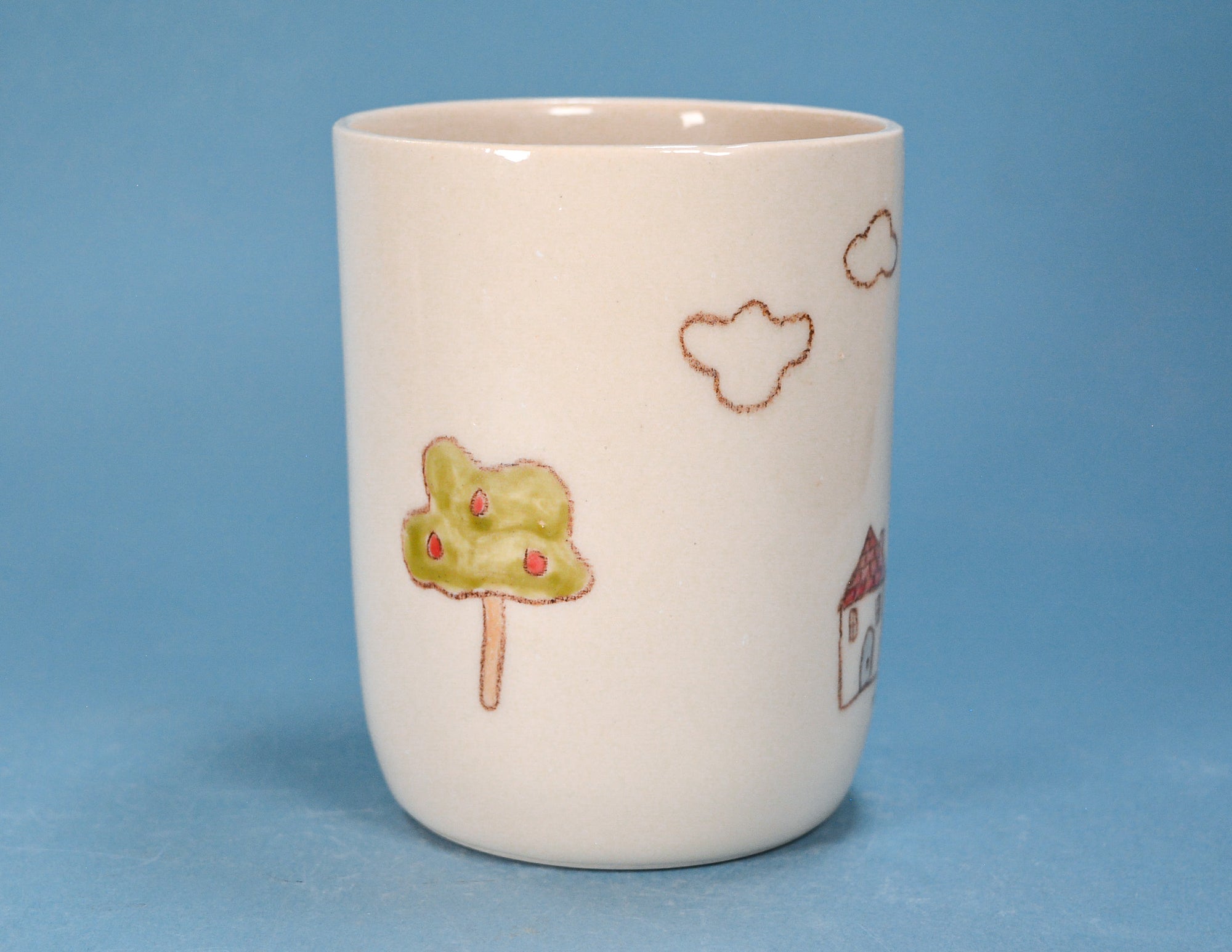 A white ceramic cup decorated with a whimsical hand-painted style illustration of a green tree with red berries, brown clouds, and a small house, set against a solid blue background.