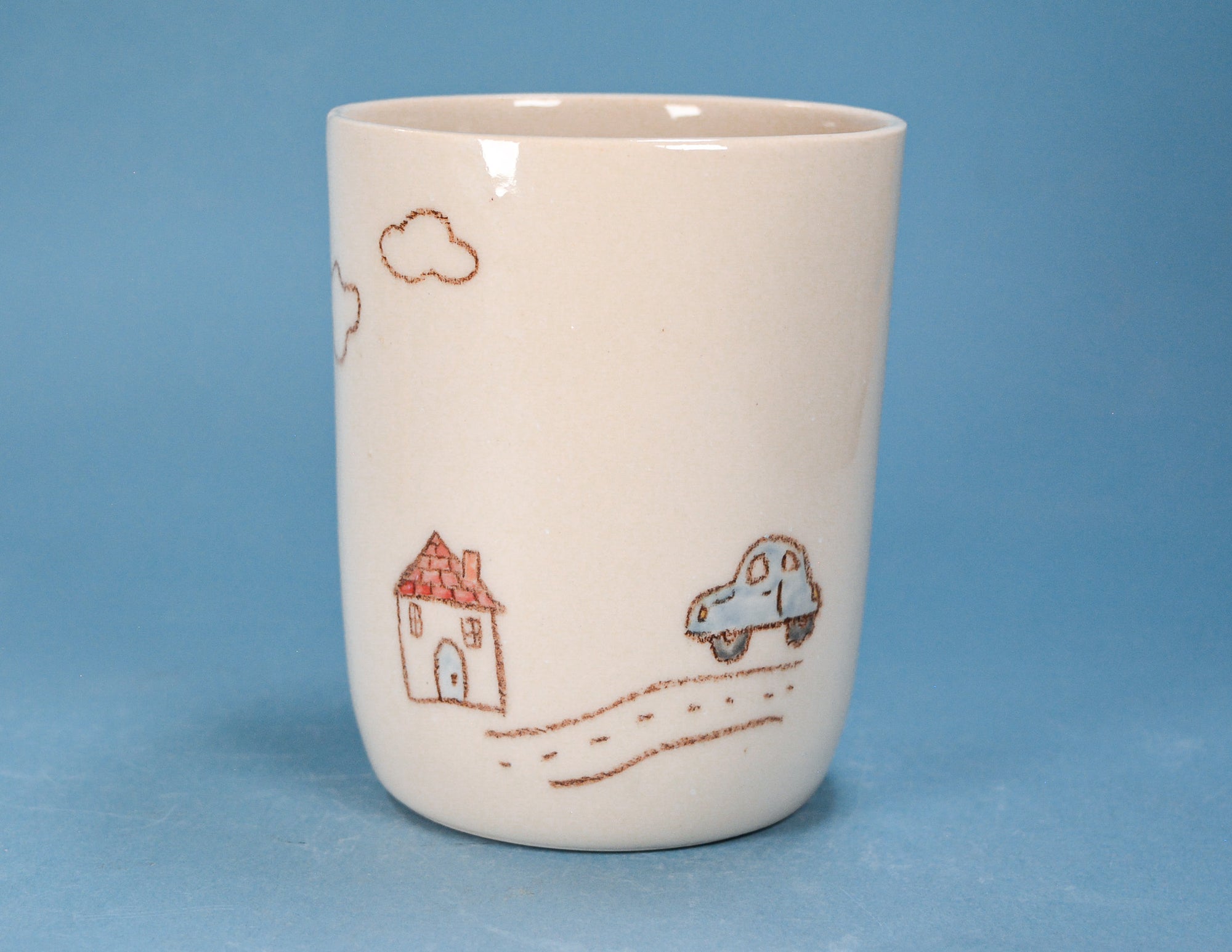 An off-white ceramic cup featuring a hand-drawn style illustration of a small house, a blue car, and clouds in brown, set against a solid blue background.
