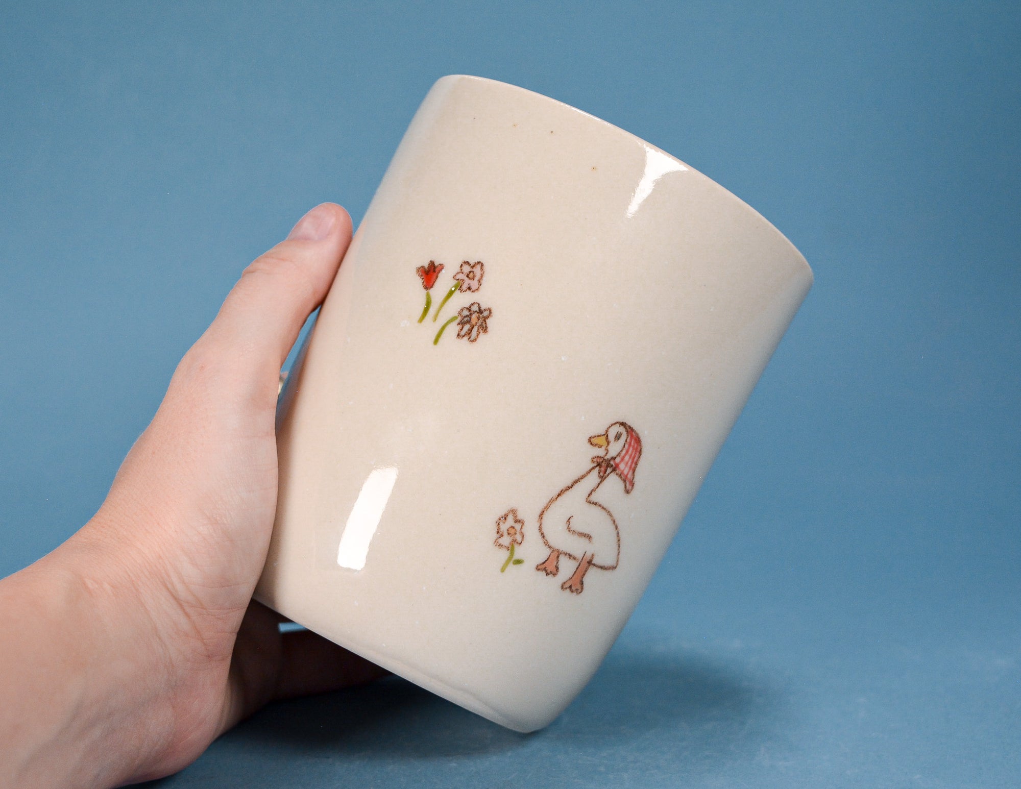 A hand holding a cream-colored ceramic mug with a glossy finish, decorated with a small line-art illustration of a goose wearing a pink hat and a cluster of flowers. The background is a solid blue studio backdrop.