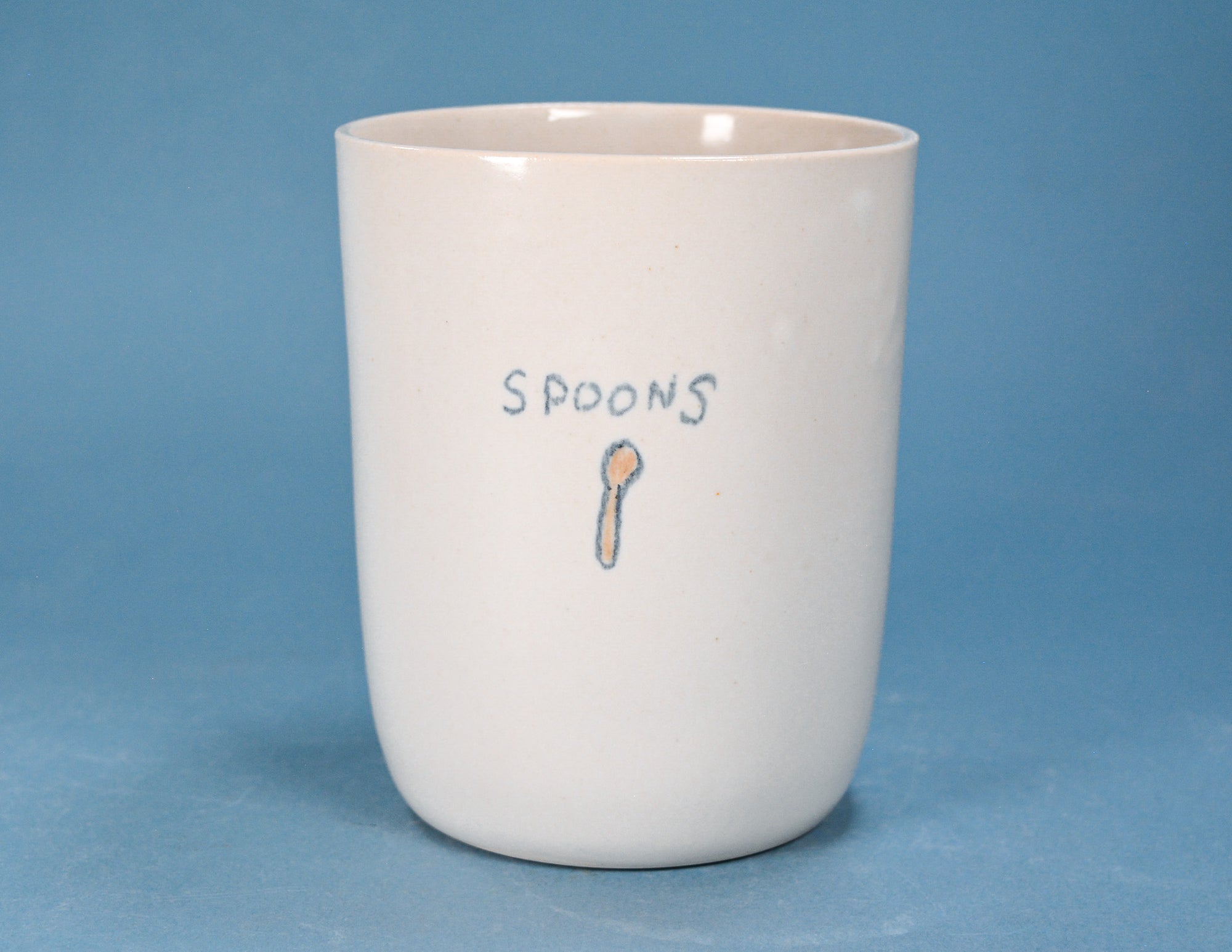 A white ceramic cup with the word 'SPOONS' and a small spoon illustration printed on the front, set against a solid blue background.