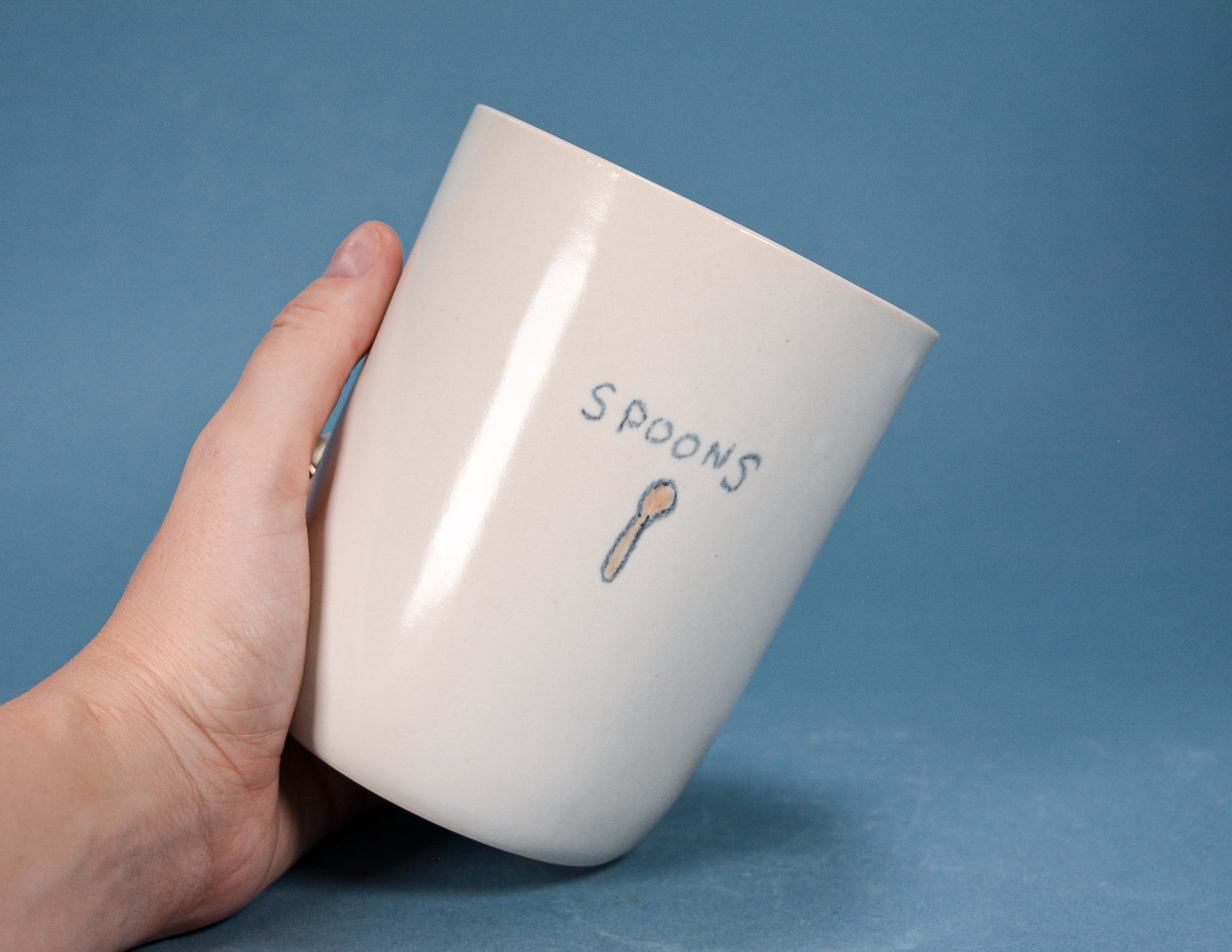 A hand holding a glossy off-white ceramic mug with the word 'SPOONS' and a small spoon illustration printed on the side, set against a solid blue background.