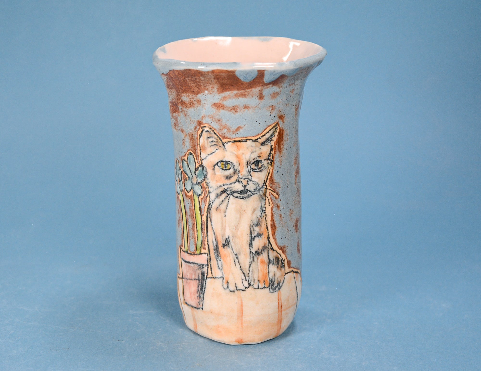 A handmade ceramic vase featuring a painted illustration of an orange tabby cat sitting next to flowers, set against a textured brown and beige glaze on a solid blue background.