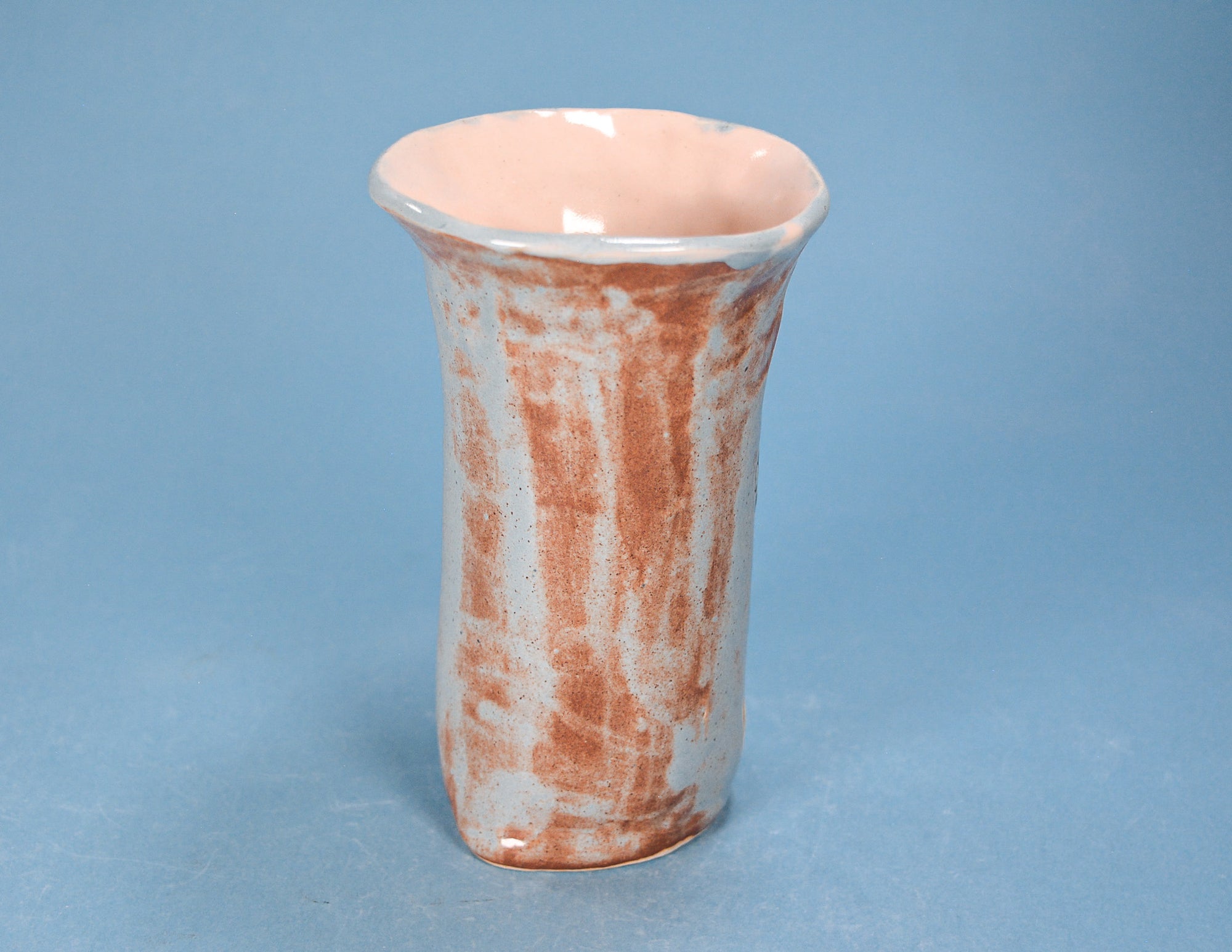 A small ceramic vase with a flared rim, featuring a textured, mottled glaze in terracotta brown and off-white, set against a solid blue background.