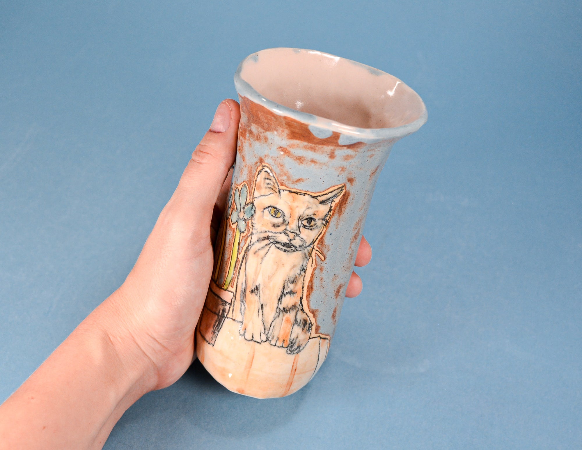A hand holds a handmade ceramic cup or vase featuring a painted cat design, set against a solid blue background.