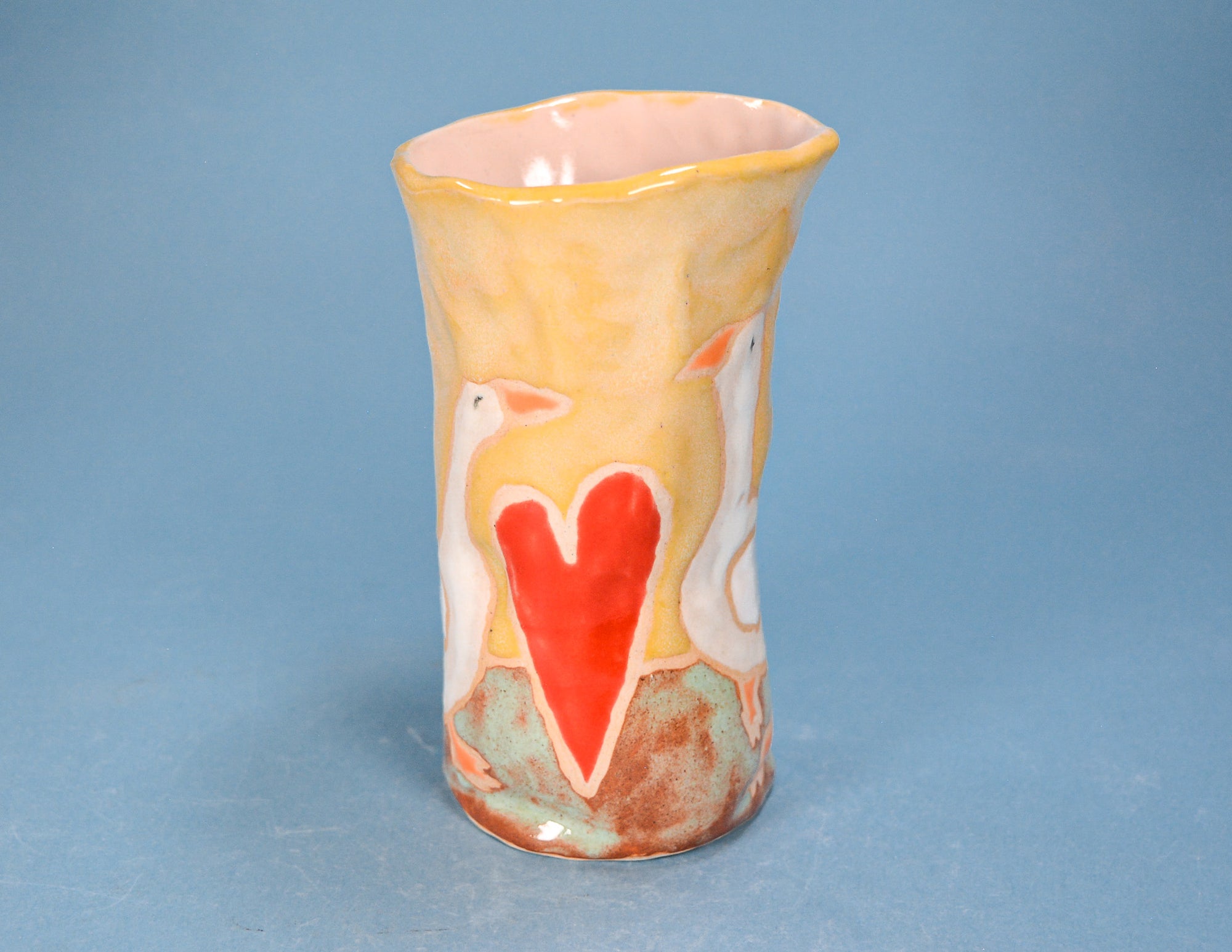 A handmade ceramic vase with a flared rim, featuring a yellow and orange marbled glaze, white bird silhouettes, and a prominent red heart on the front, set against a solid blue background.