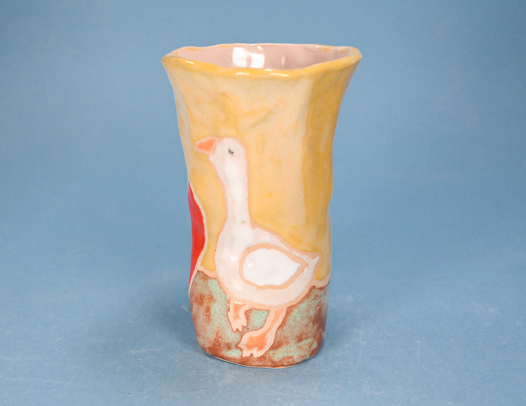 A handmade ceramic vase featuring a painted white goose on a textured yellow and orange body, set against a solid blue background.