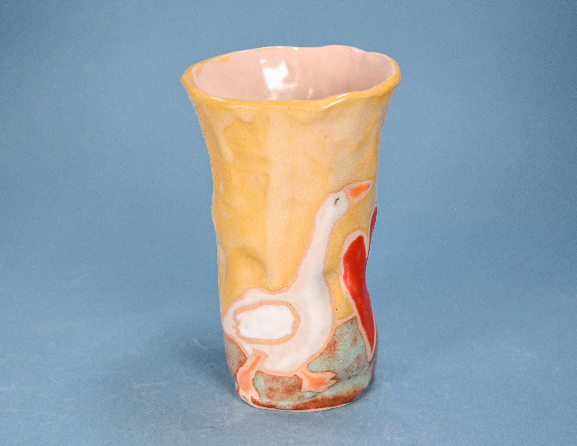A handmade ceramic cup with a flared rim, featuring a yellow and orange marbled glaze with a white goose and red graphic design on the side, set against a solid blue background.