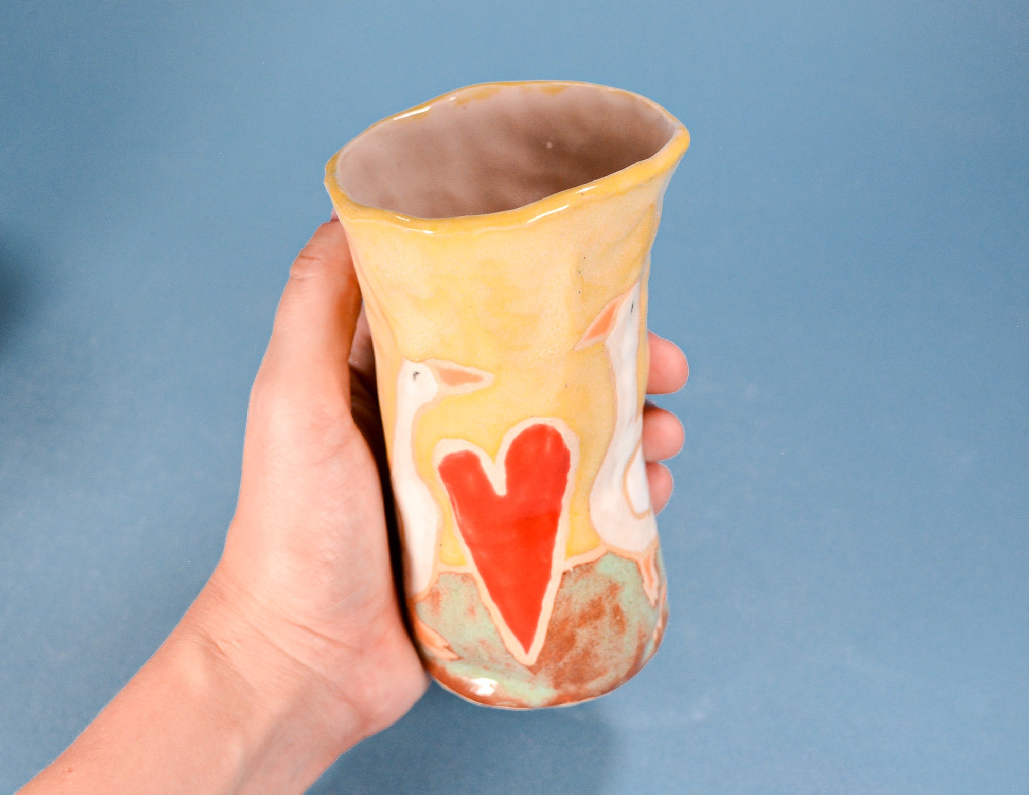 A hand holding a handmade ceramic cup with a yellow and orange glaze, featuring a red heart and white swan design, set against a solid blue background.