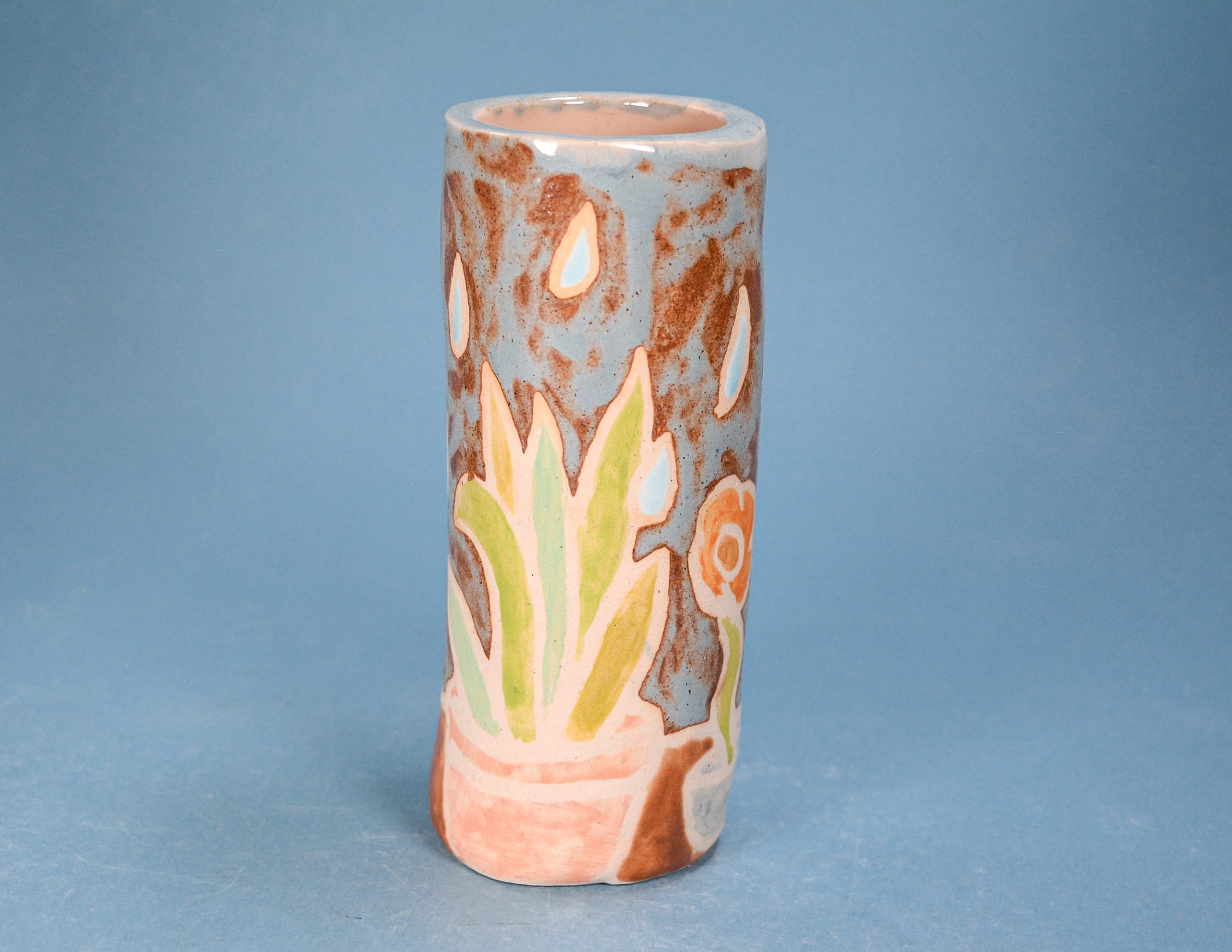 A handmade ceramic cylindrical vase with a rustic design, featuring a light blue-gray background with brown speckles, a green succulent plant, and peach-colored accents, set against a solid blue background.